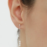 Close-up of a womans ear wearing the Eden Trio Pearl Drop Earrings in Silver, featuring a small stud and three white pearls dangling vertically from a thin silver chain. These elegant earrings stand out against a plain, neutral background.
