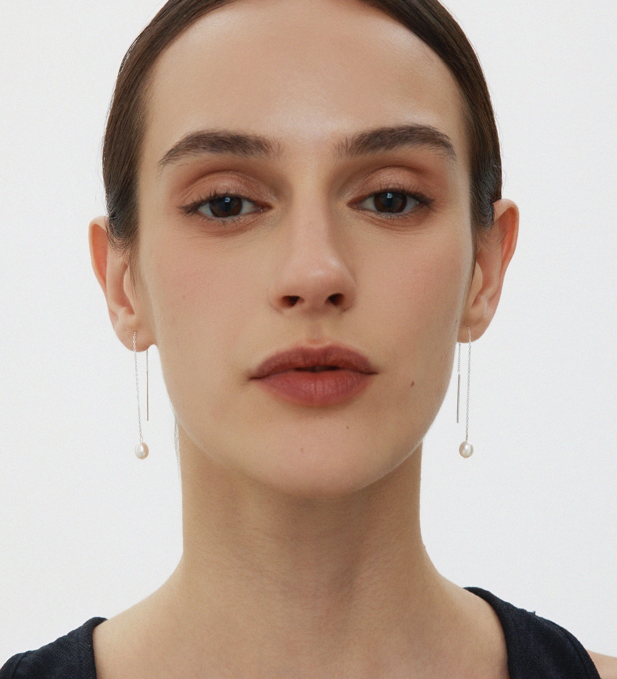 A woman with straight dark hair pulled back wears Eira Pearl Drop Threader Earrings in Silver and a sleeveless black top, facing forward against a plain white background.