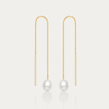 The Eira Pearl Drop Threader Earrings in Gold feature delicate chains ending with a single round white pearl, offering a minimalist and elegant look against a plain white background.