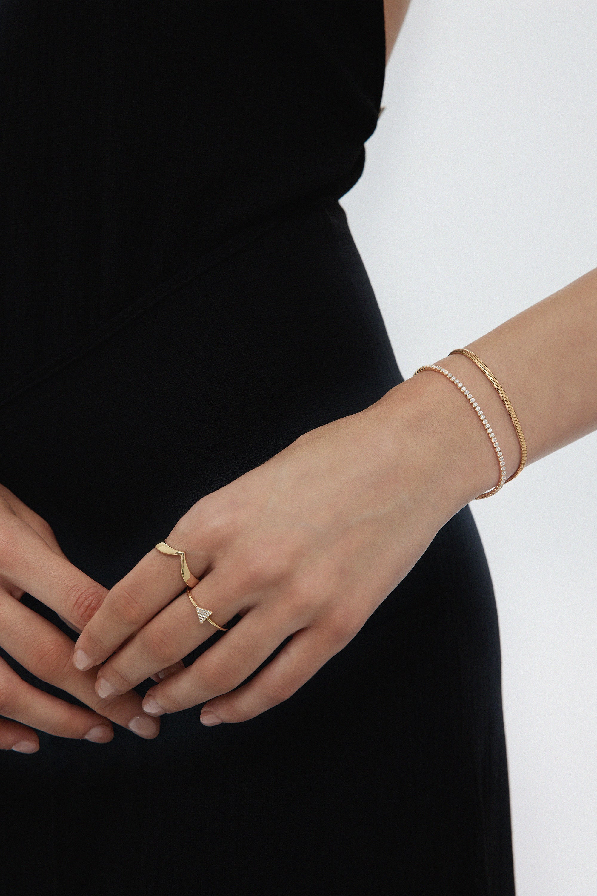 A person in a black dress shows their left hand adorned with two gold rings and a stylish bracelet stack, featuring the Estelle Herringbone Chain Bracelet in Gold alongside delicate pieces, with fingers gently resting on their right hand.