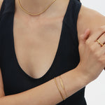 A woman in a sleeveless black top displays gold jewelry: a 3mm chain necklace, the Estelle Herringbone Chain Bracelet in Gold stacked with others, and a ring; her arms are gently crossed, her face partially out of frame.