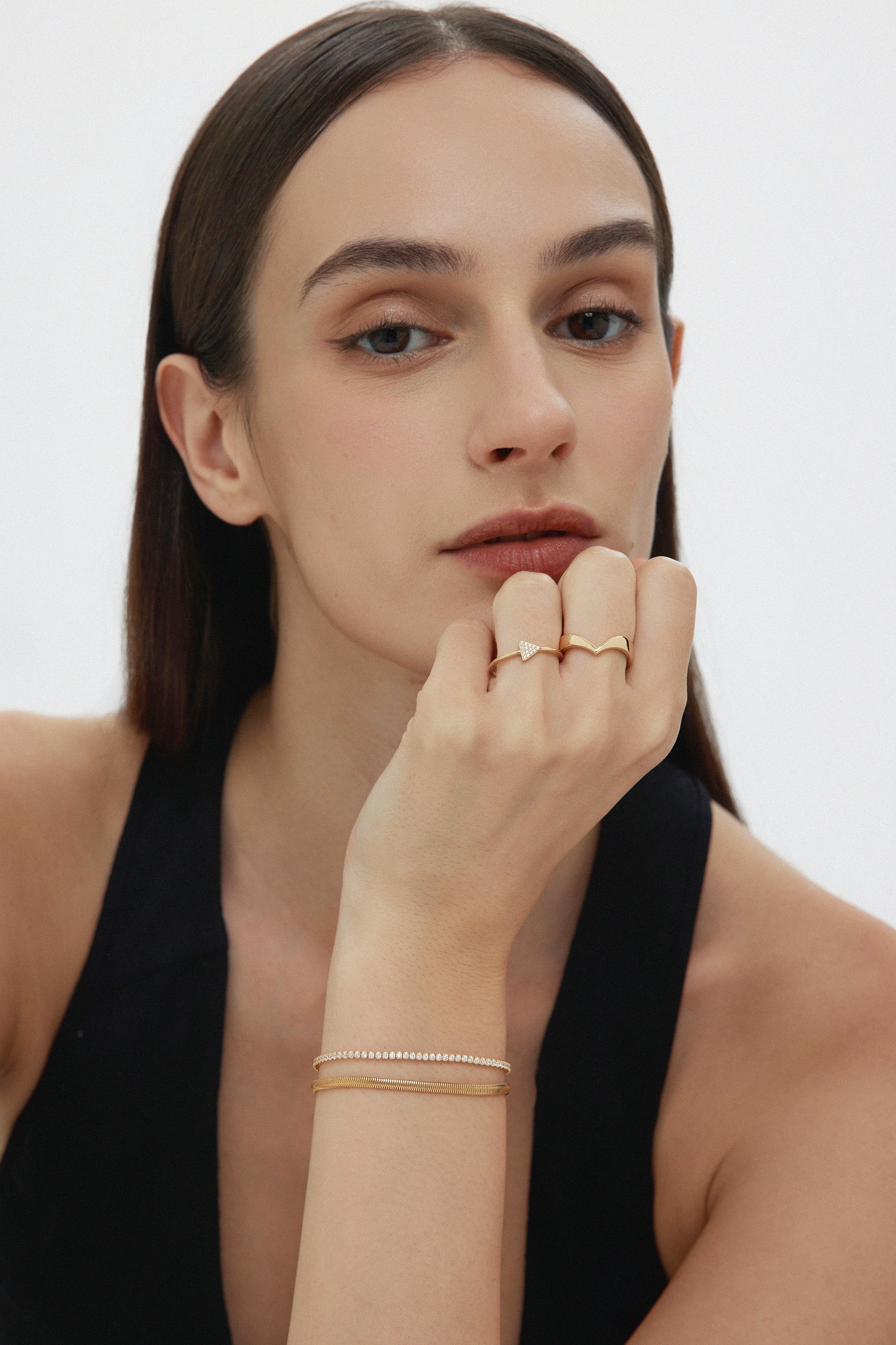 A woman with straight brown hair and light skin wears a black sleeveless top, gold rings, and the Estelle Herringbone Chain Bracelet in Gold as part of her chic bracelet stack, resting her chin on her hand and gazing confidently at the camera.