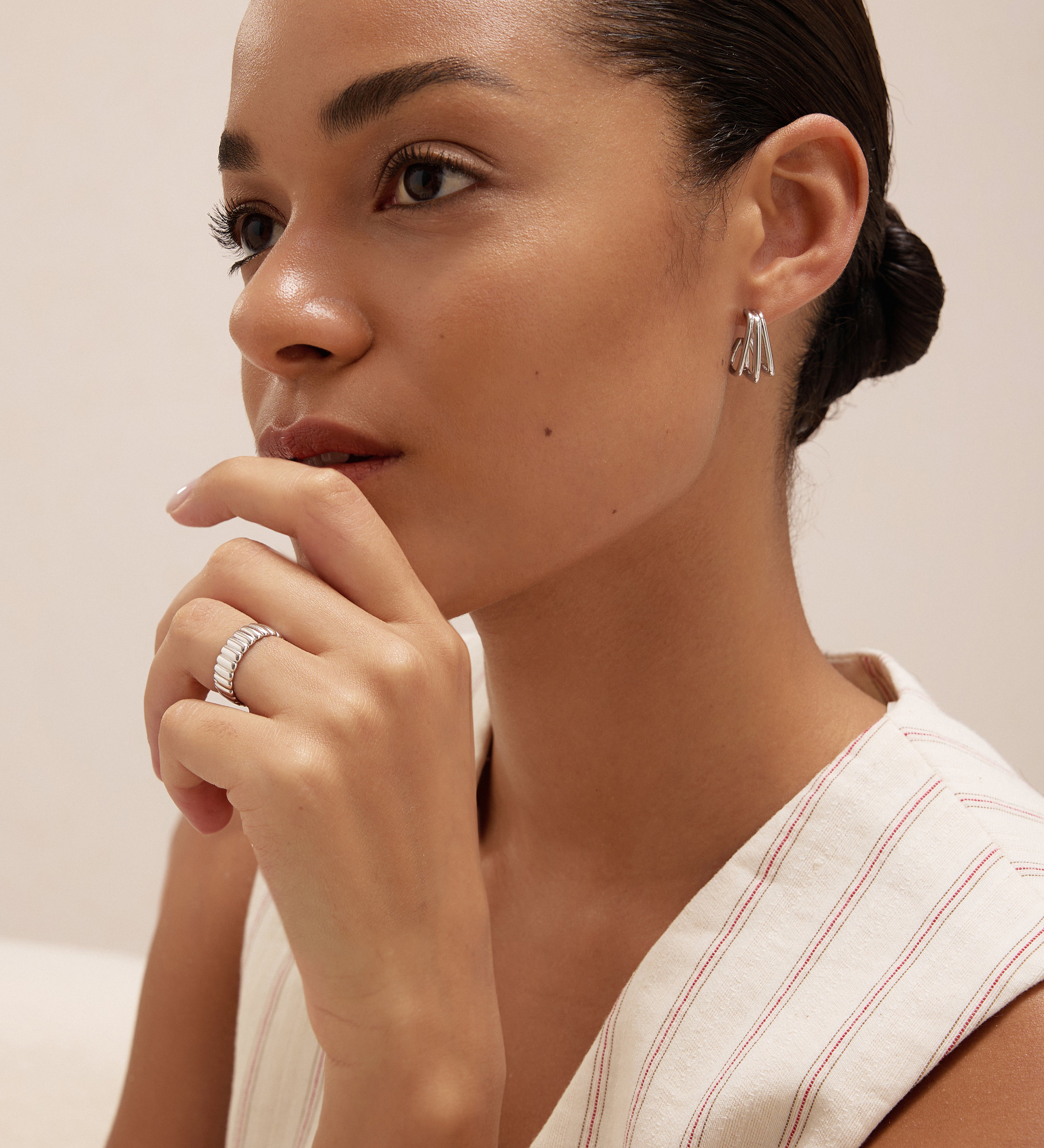 A woman with her hair pulled back wears the Esther Square Hoop Stud Earrings in Silver and a matching ring, resting her hand near her chin. She is dressed in a light sleeveless striped top, gazing thoughtfully into the distance.