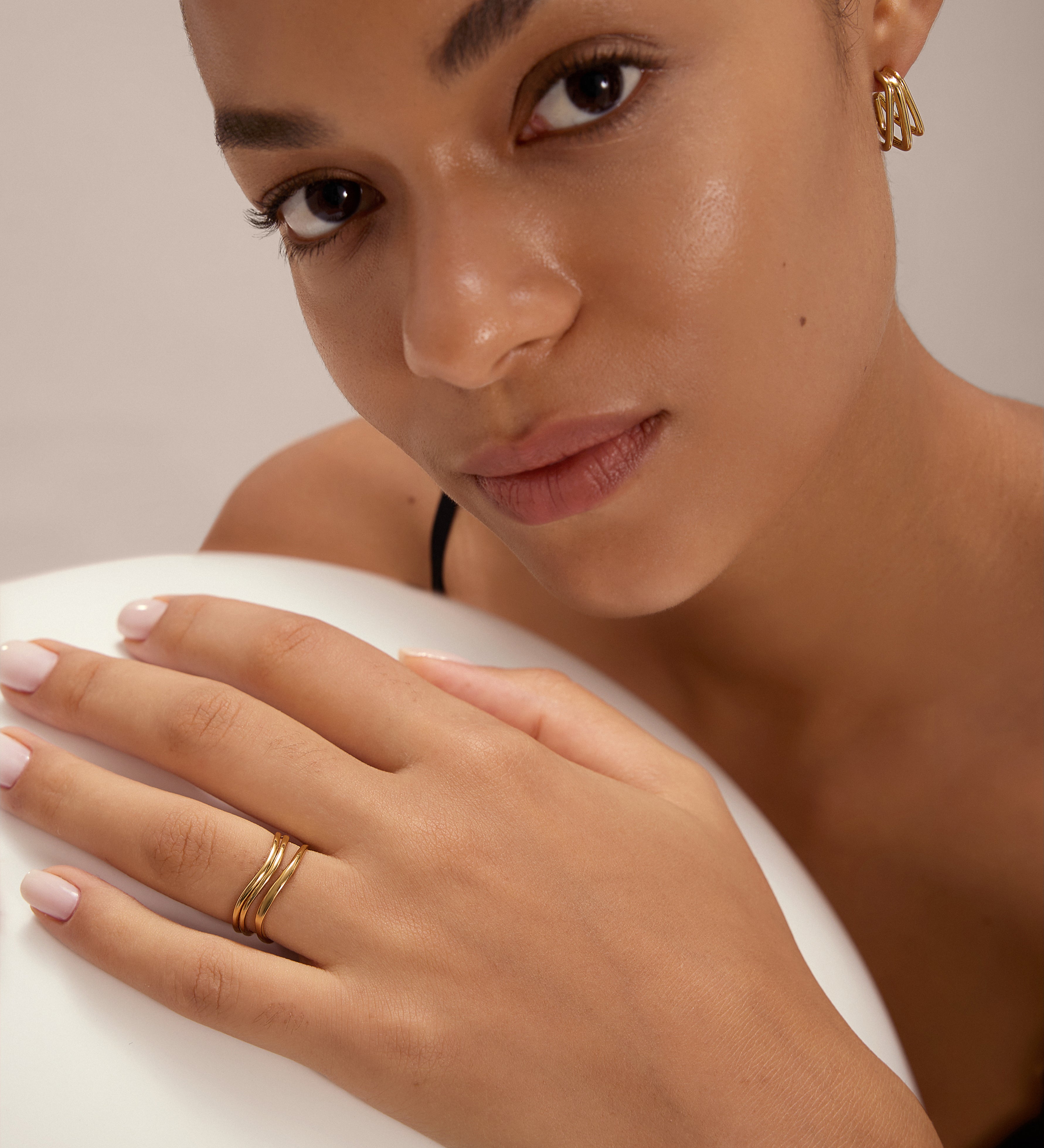 A woman with glowing skin and natural makeup wears Esther Square Hoop Stud Earrings in Gold. She rests her manicured hand on a white surface, gazing softly at the camera.