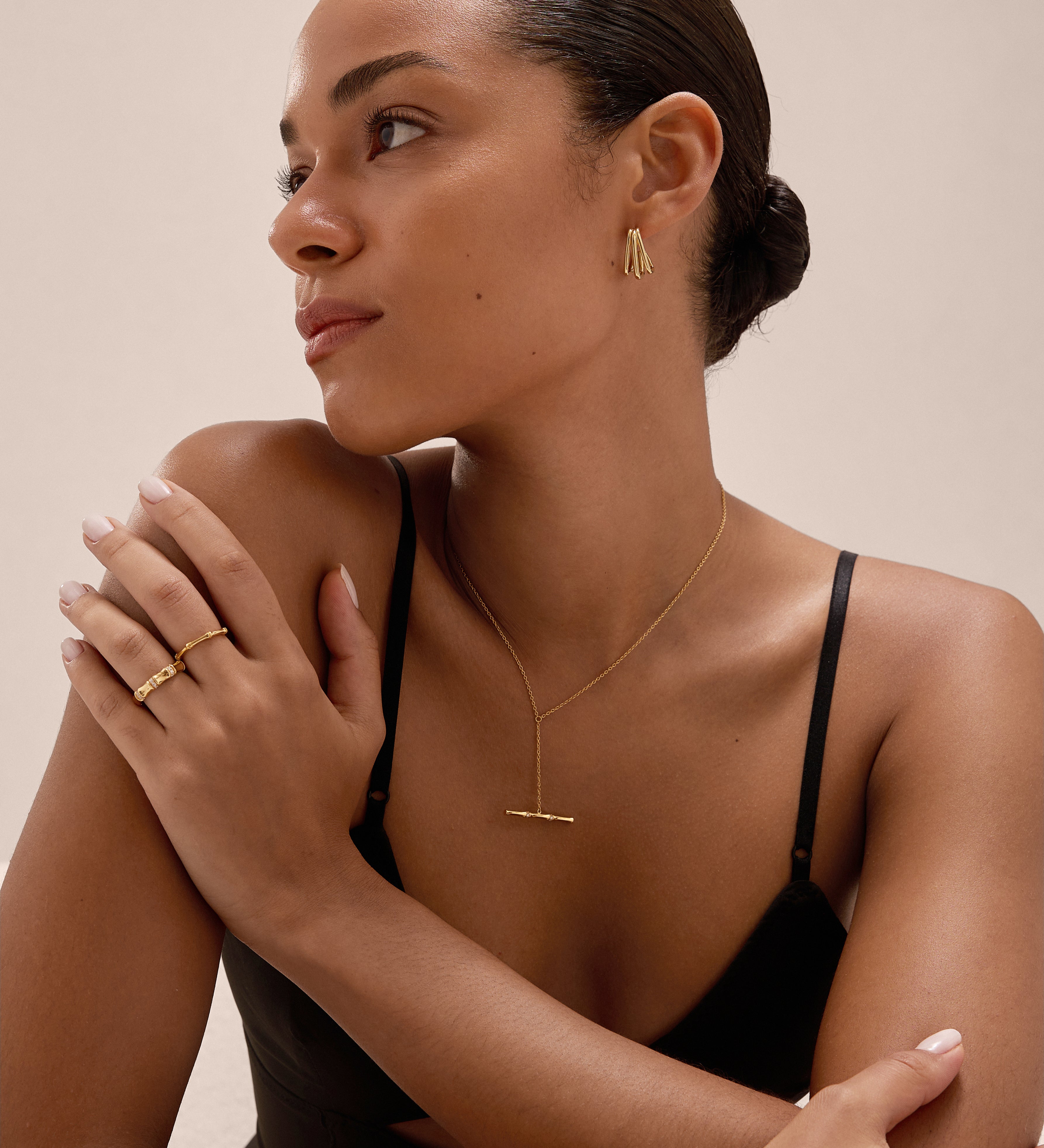 A woman with smooth, pulled-back hair wears a black strappy top and gold jewelry, including the Esther Square Hoop Stud Earrings in Gold, as she looks to the side with her arms gently crossed against a neutral background.