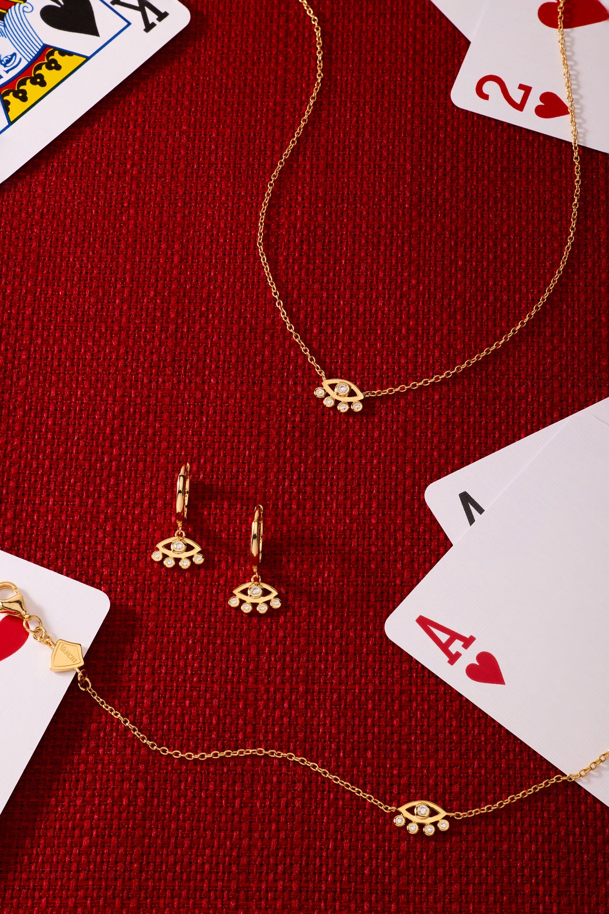 Silver Defender Evil Eye Necklace arranged with matching gold jewelry—bracelet, earrings, and charms—on a red textured surface, surrounded by playing cards like the ace of hearts and king.