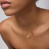 A woman in a strapless brown top showcases the Defender Evil Eye Necklace in Gold, 18k gold plated with a small cubic zirconia pendant, against a neutral background. Her face is partially visible, emphasizing her lips and jawline.