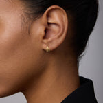 Close-up of a woman wearing Flame Stud Earrings in Gold—small, 18k gold-plated multi-bar studs. Her dark hair is pulled back, she's in a black collared top, and the background is soft and neutral.