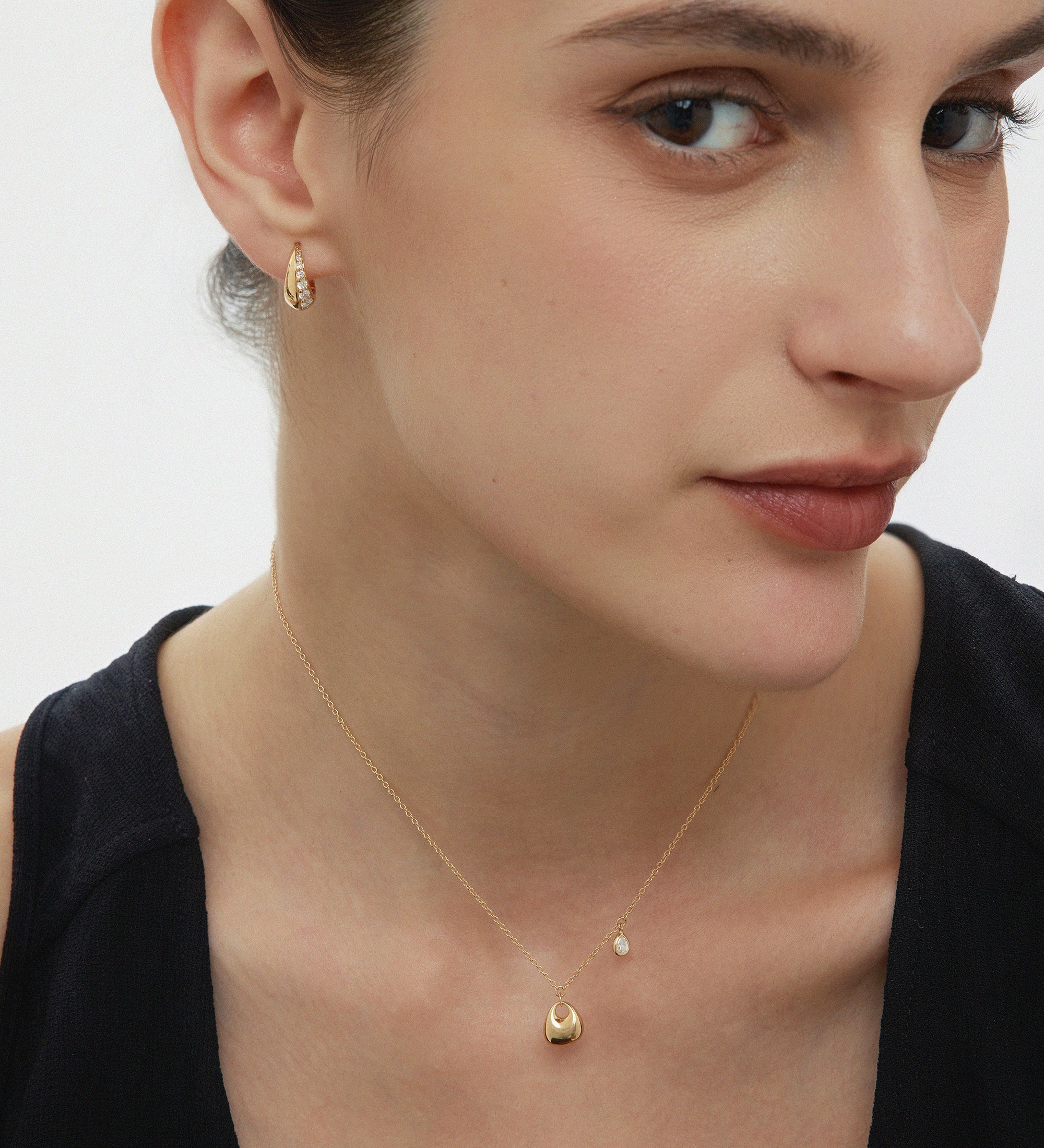 A woman with fair skin and brown hair pulled back wears a black top, a delicate gold necklace, and Georgie Crystal Knife Edge Huggie Hoop Earrings in Gold. She gazes softly to the side with a neutral expression.