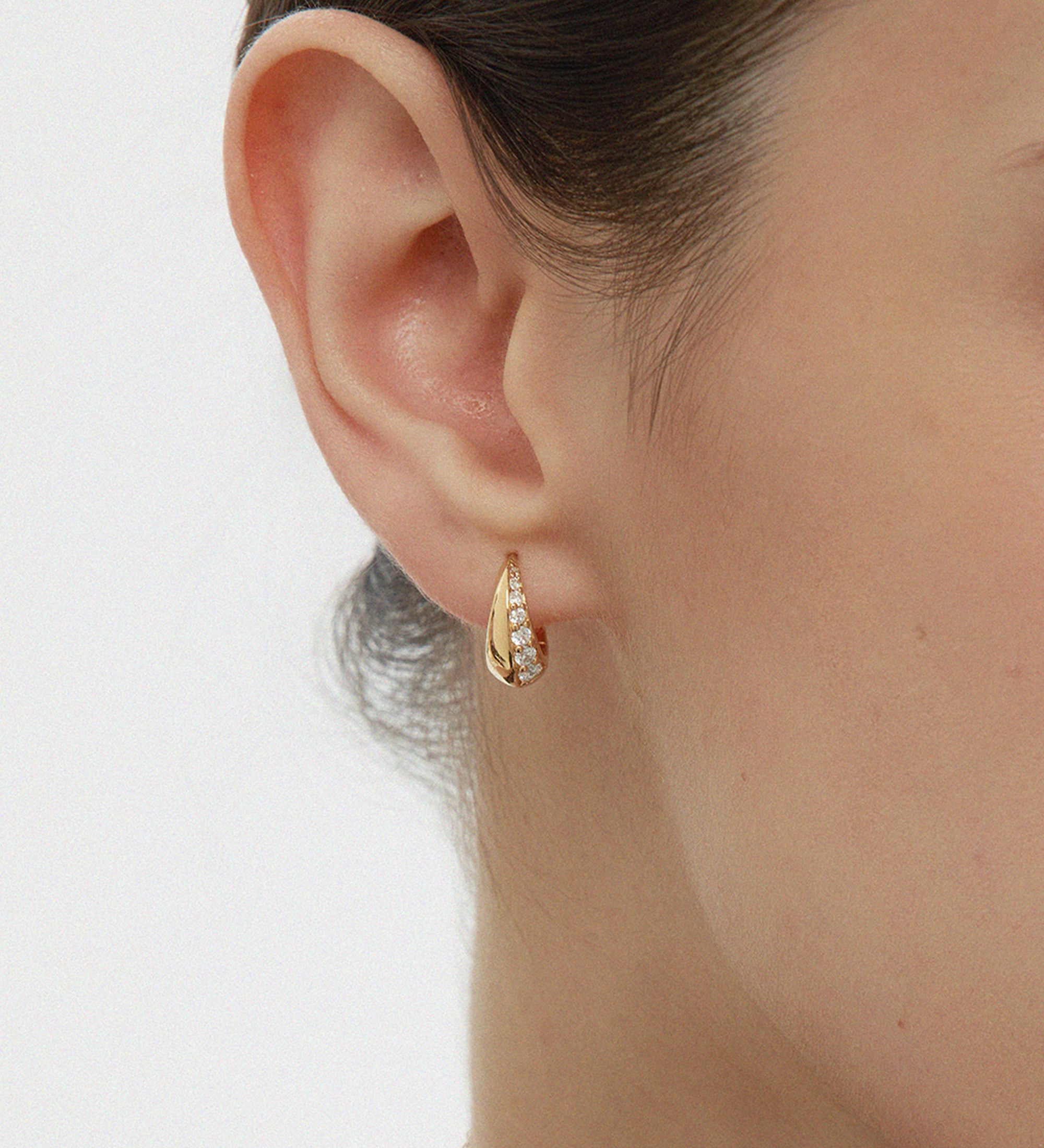 Close-up of a woman’s ear wearing the Georgie Crystal Knife Edge Huggie Hoop Earrings in Gold, featuring embedded cubic zircon stones. Her hair is tied back and a delicate gold chain necklace is partially visible.