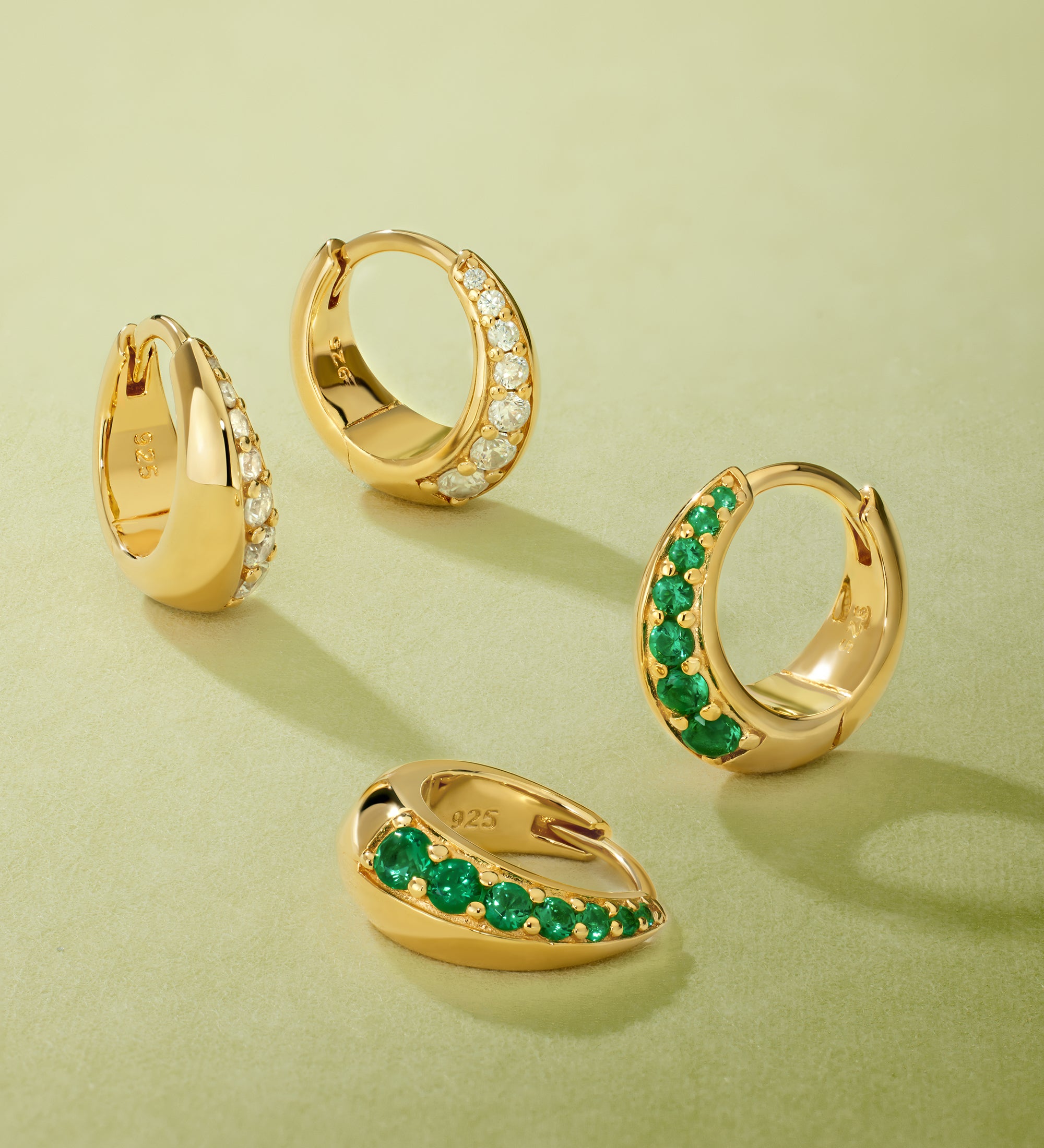 Four Georgie Crystal Knife Edge Huggie Hoop Earrings in Gold rest on a light green surface; two feature clear crystals, and two have green stones, each arranged at varying angles.