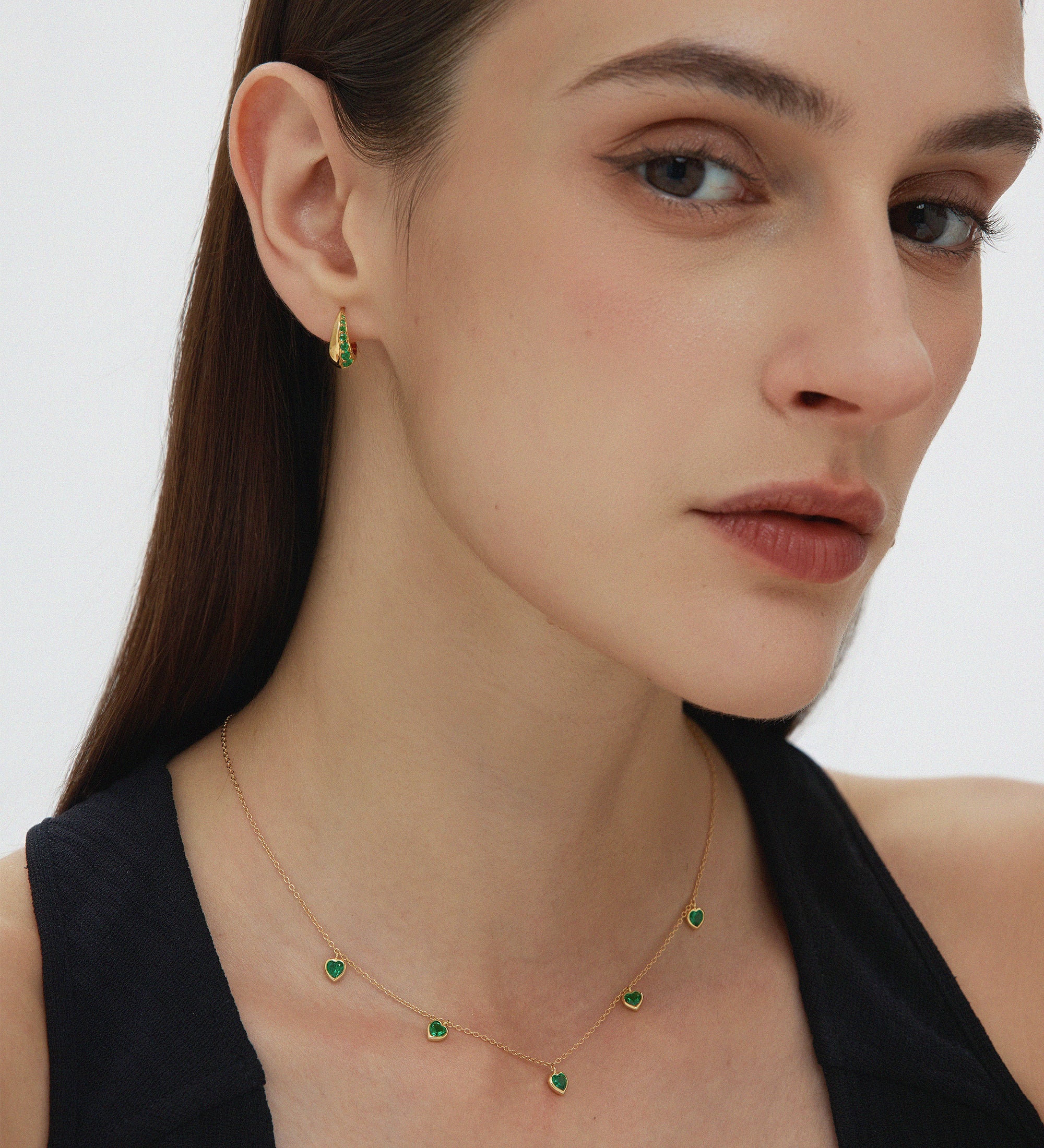 A woman with straight brown hair wears a black top, a gold necklace with green stones, and Georgie Crystal Knife Edge Huggie Hoop Earrings in Green, posing against a plain light background.