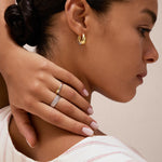 A woman with neat dark hair touches her neck, showcasing Gia Reversible 2 Tone Chubby Hoop Earrings and two rings—one with white stones. She wears a light striped top and has manicured pale pink nails.