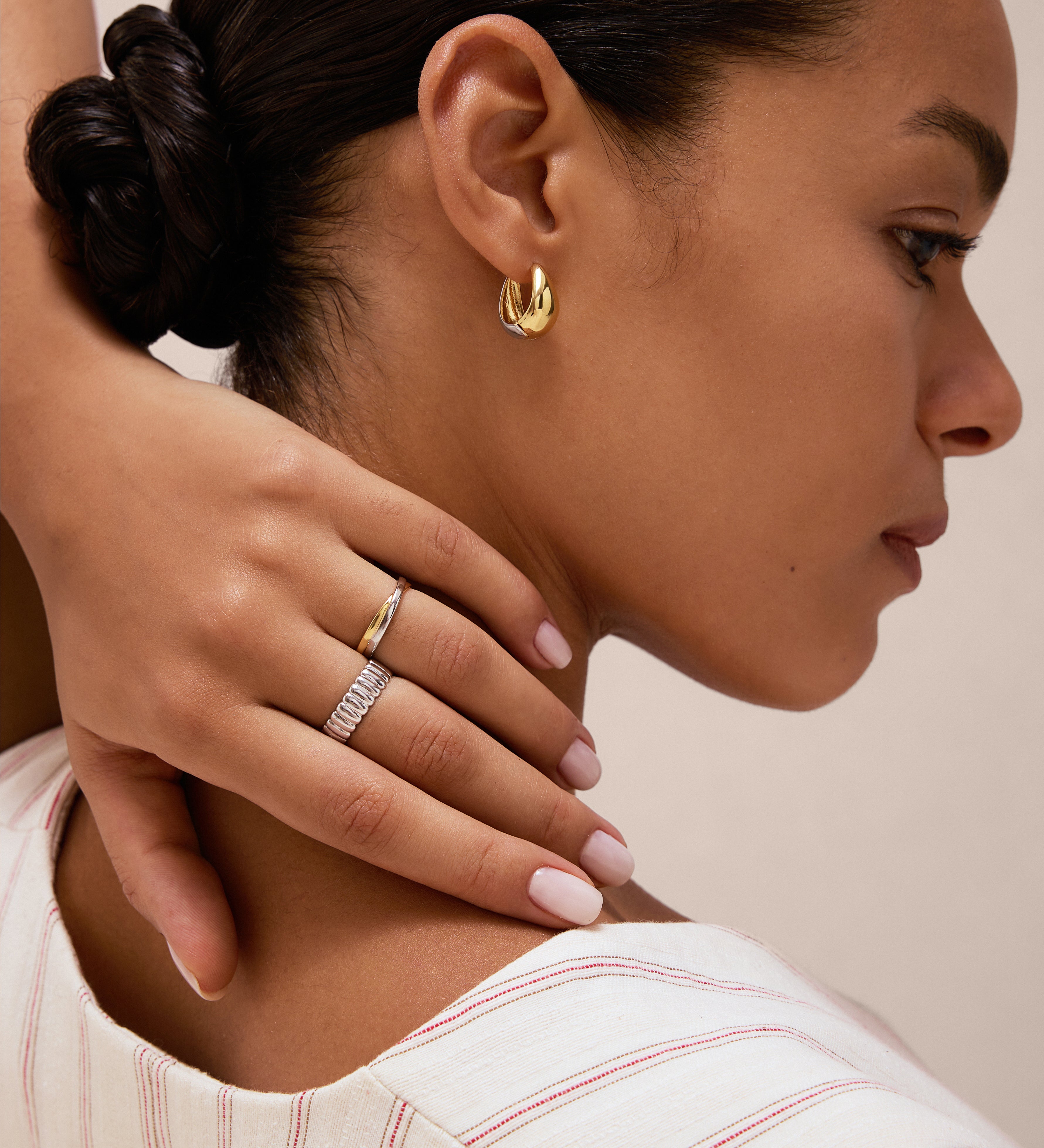 A woman with neat dark hair touches her neck, showcasing Gia Reversible 2 Tone Chubby Hoop Earrings and two rings—one with white stones. She wears a light striped top and has manicured pale pink nails.