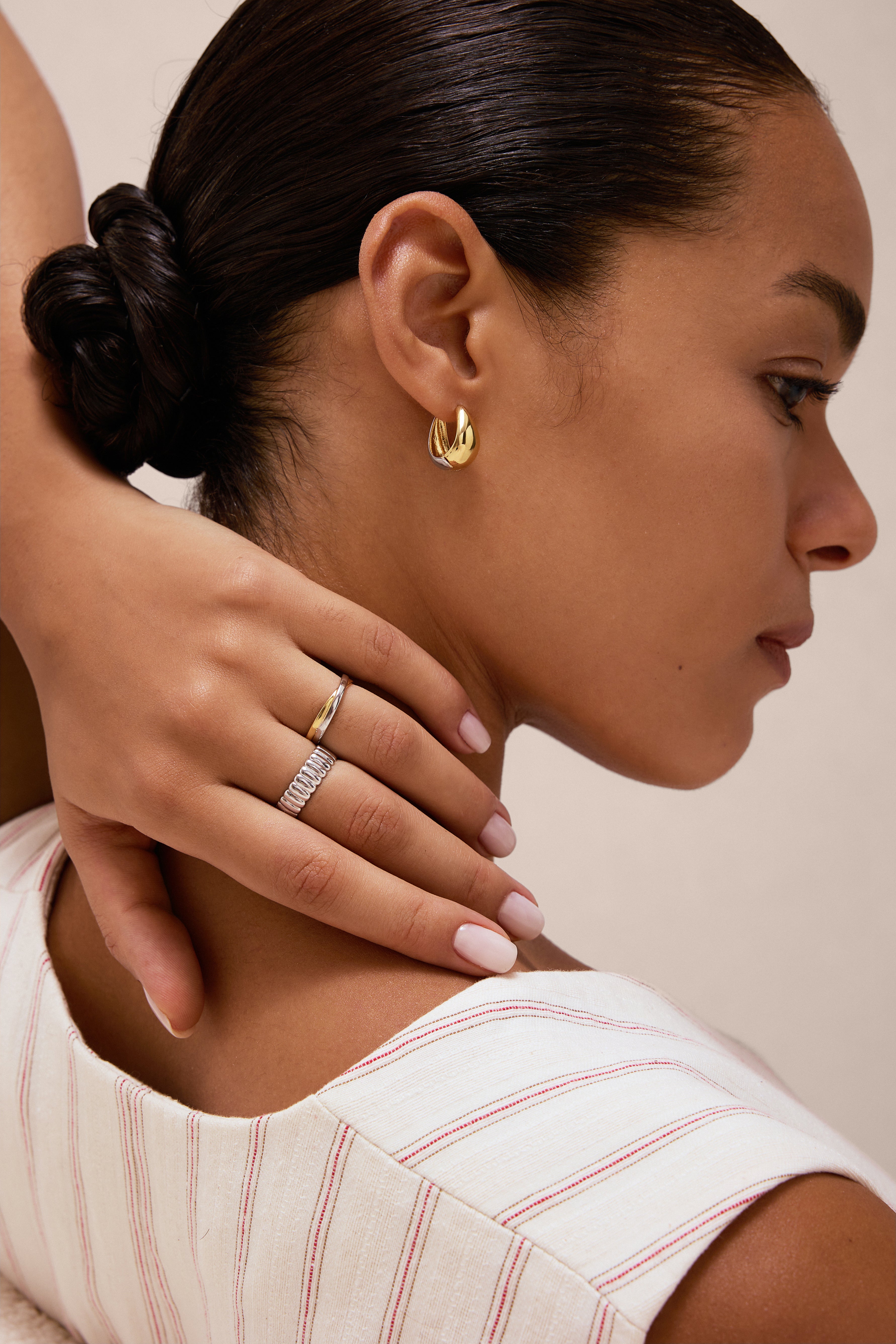 A woman with neat dark hair touches her neck, showcasing Gia Reversible 2 Tone Chubby Hoop Earrings and two rings—one with white stones. She wears a light striped top and has manicured pale pink nails.