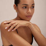 A woman with smooth skin and slicked-back hair rests her chin on her hand, wearing the Gia Reversible 2 Tone Chubby Hoop Earrings in 18k gold and sterling silver. She has light pink nails, a black top, and is posed against a neutral background.