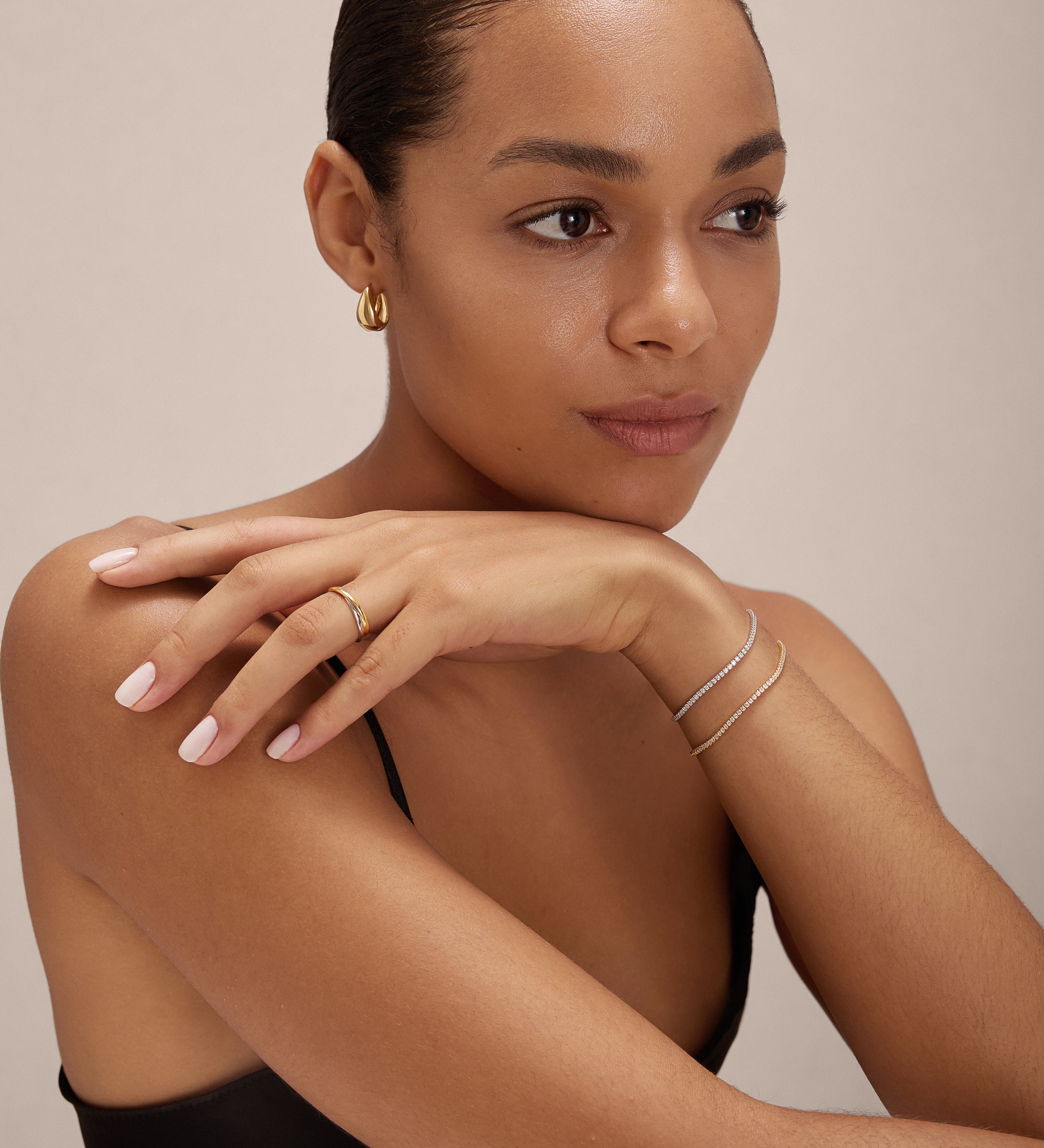A woman with smooth skin and slicked-back hair rests her chin on her hand, wearing the Gia Reversible 2 Tone Chubby Hoop Earrings in 18k gold and sterling silver. She has light pink nails, a black top, and is posed against a neutral background.