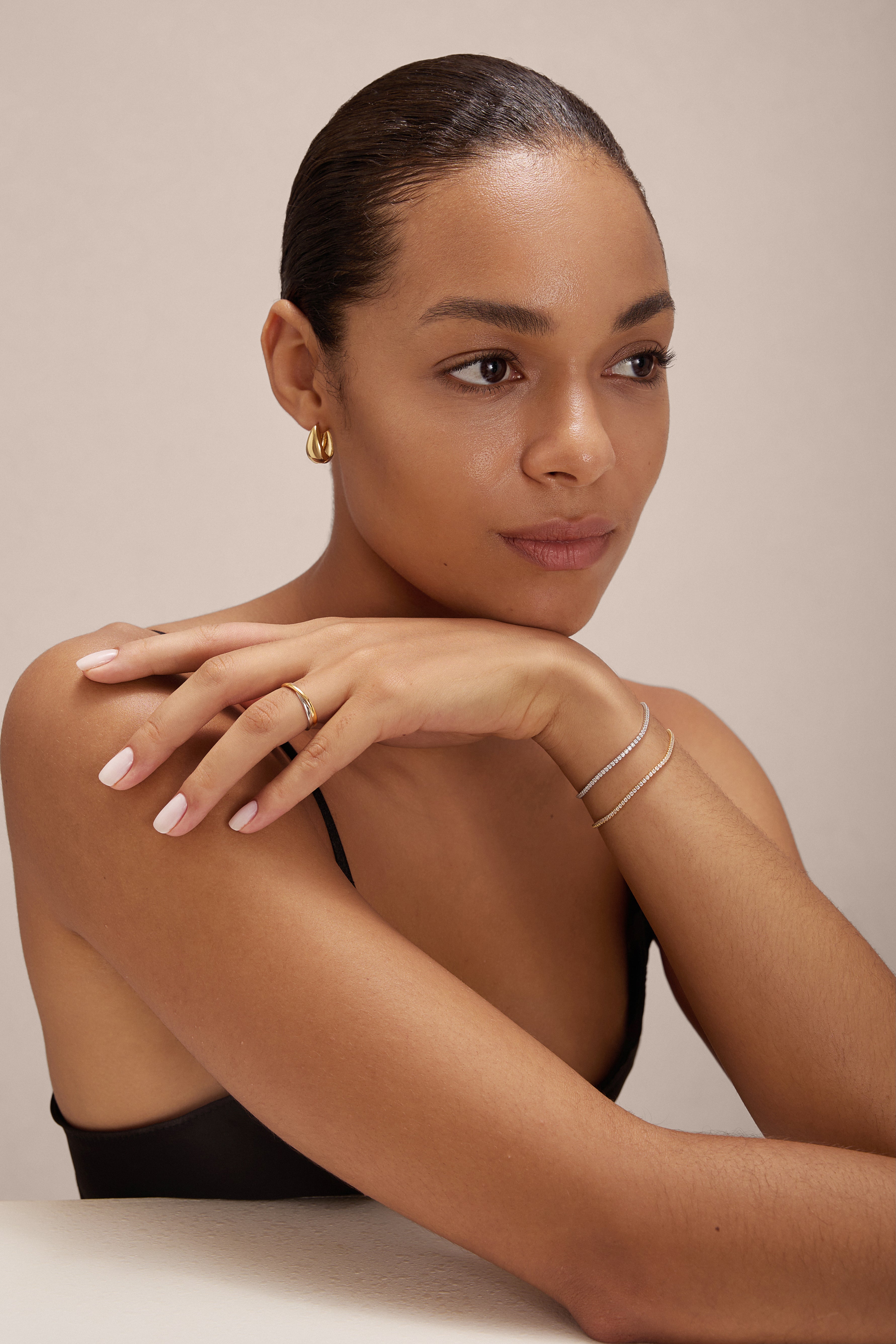 A woman with smooth skin and slicked-back hair rests her chin on her hand, wearing the Gia Reversible 2 Tone Chubby Hoop Earrings in 18k gold and sterling silver. She has light pink nails, a black top, and is posed against a neutral background.