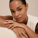 A woman with sleek, pulled-back hair rests on a cream surface, wearing Gia Reversible 2 Tone Chubby Hoop Earrings and rings. Her nails are neatly manicured as she gazes calmly at the camera.