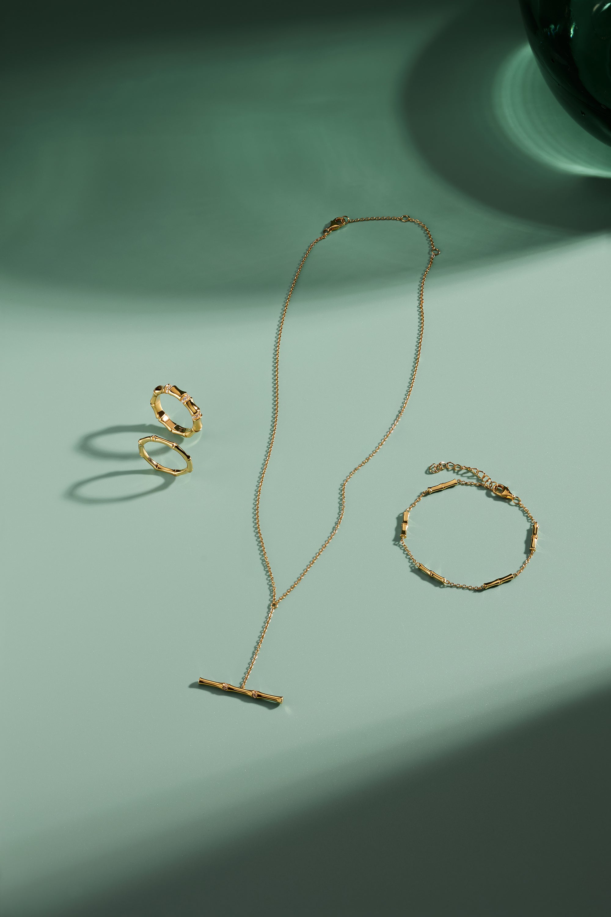 A delicate gold jewelry set arranged on a mint green surface, featuring a ring, bracelet, and the Bamboo Chain Y-Bar Lariat Necklace in Gold, each piece casting soft shadows.