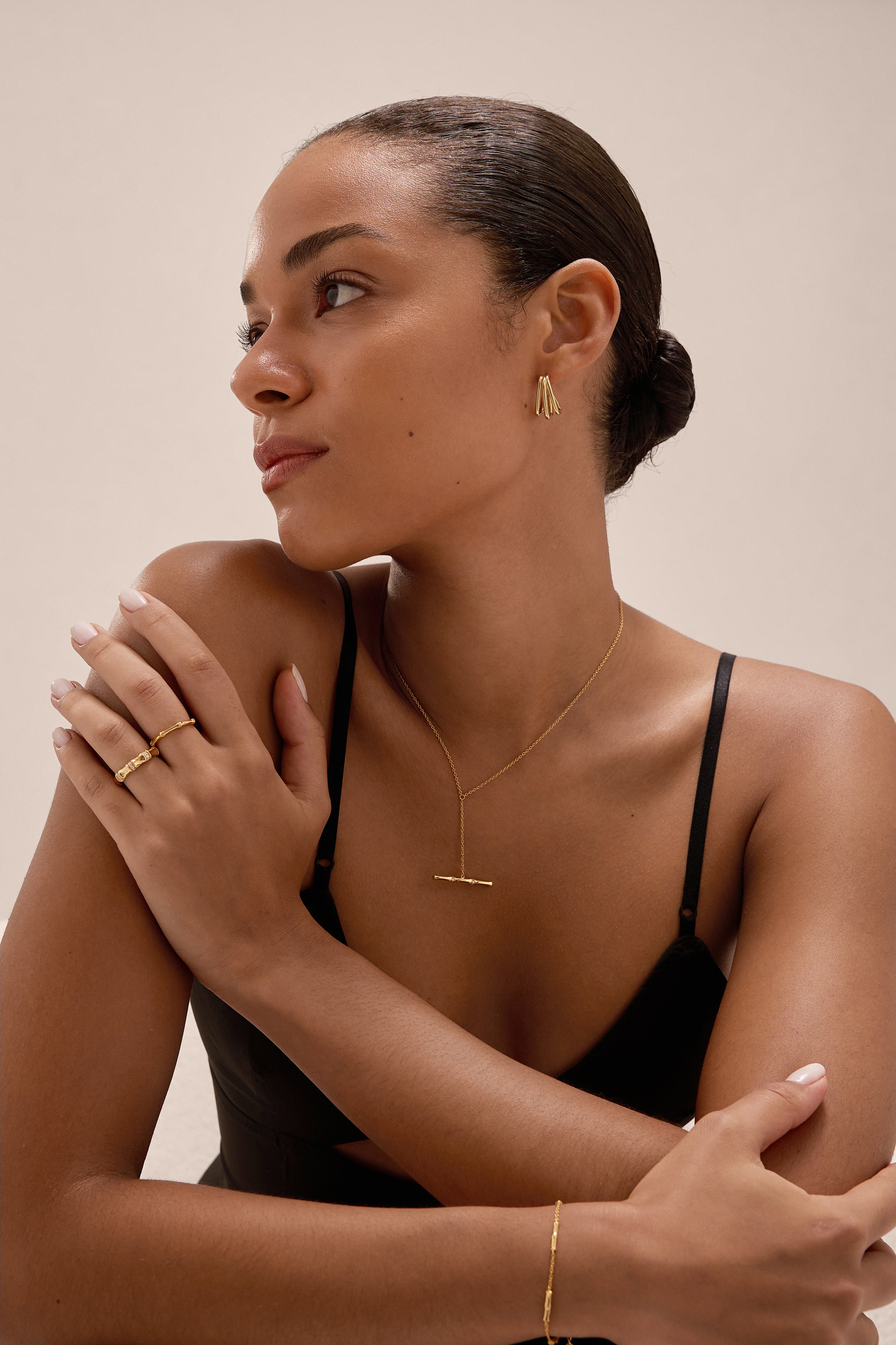 A woman with her hair in a neat bun wears minimalist gold jewelry, including the Bamboo Ring in Gold, earrings, a necklace, and bracelet. In a black spaghetti strap top, she poses with arms crossed, looking thoughtfully to the side.