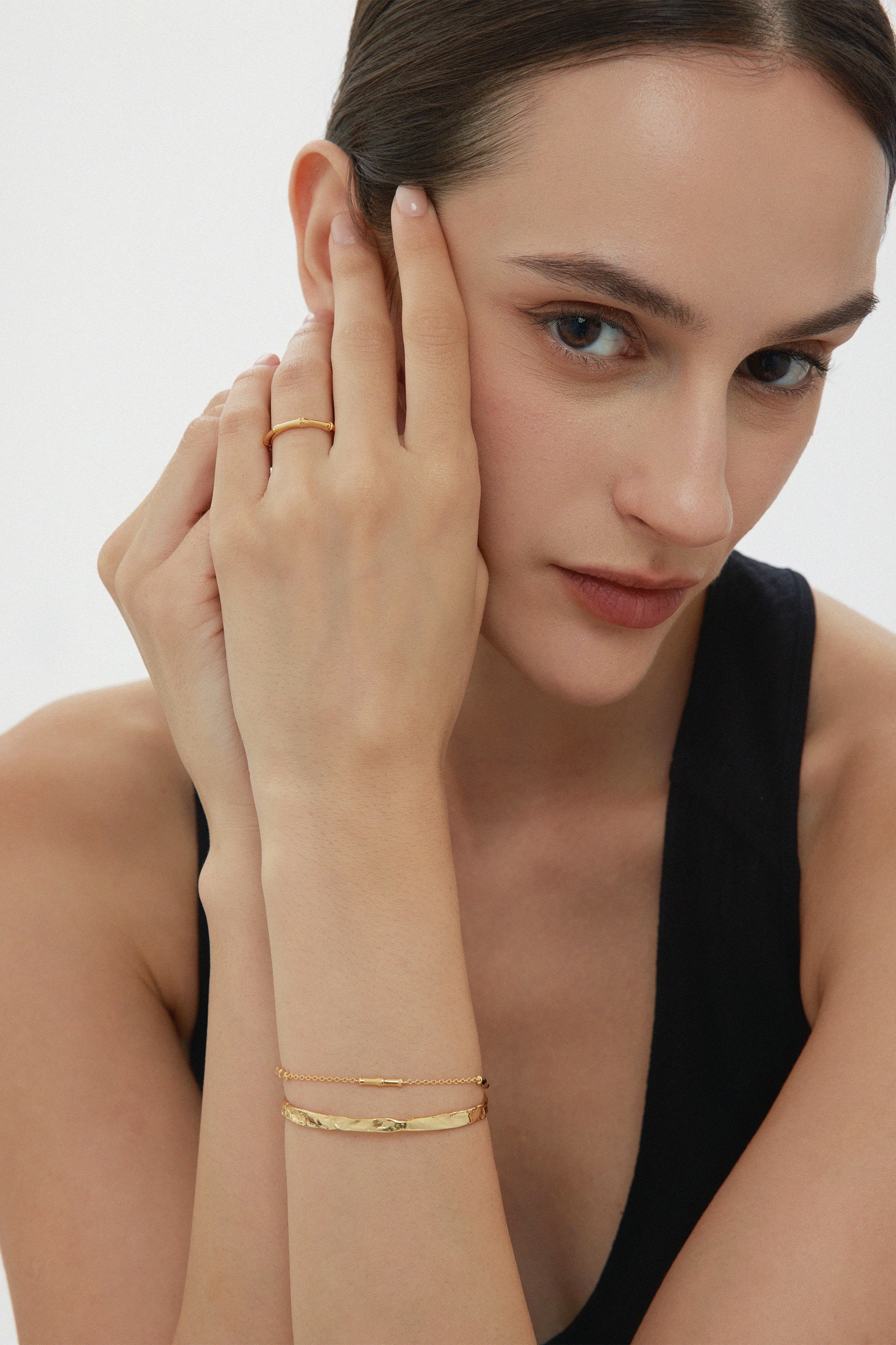 A woman with dark hair pulled back wears a sleeveless black top and minimalist gold jewelry, including the Bamboo Ring in Gold, with her hand gently touching her face as she looks at the camera.