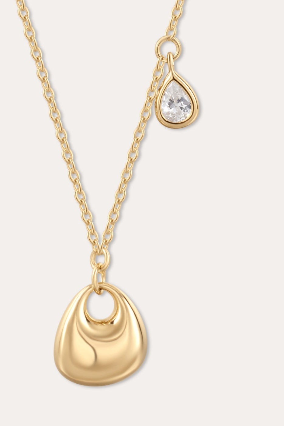 The Nora Double Pendant Necklace in Gold features a sculptural pendant paired with a small, teardrop-shaped cubic zirconia charm suspended just above it.