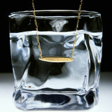 The Viva La Vida Bar Chain Necklace in Gold hangs over the rim of a clear glass filled with water, set against a white and black background, and features an adjustable figaro chain for the perfect fit.
