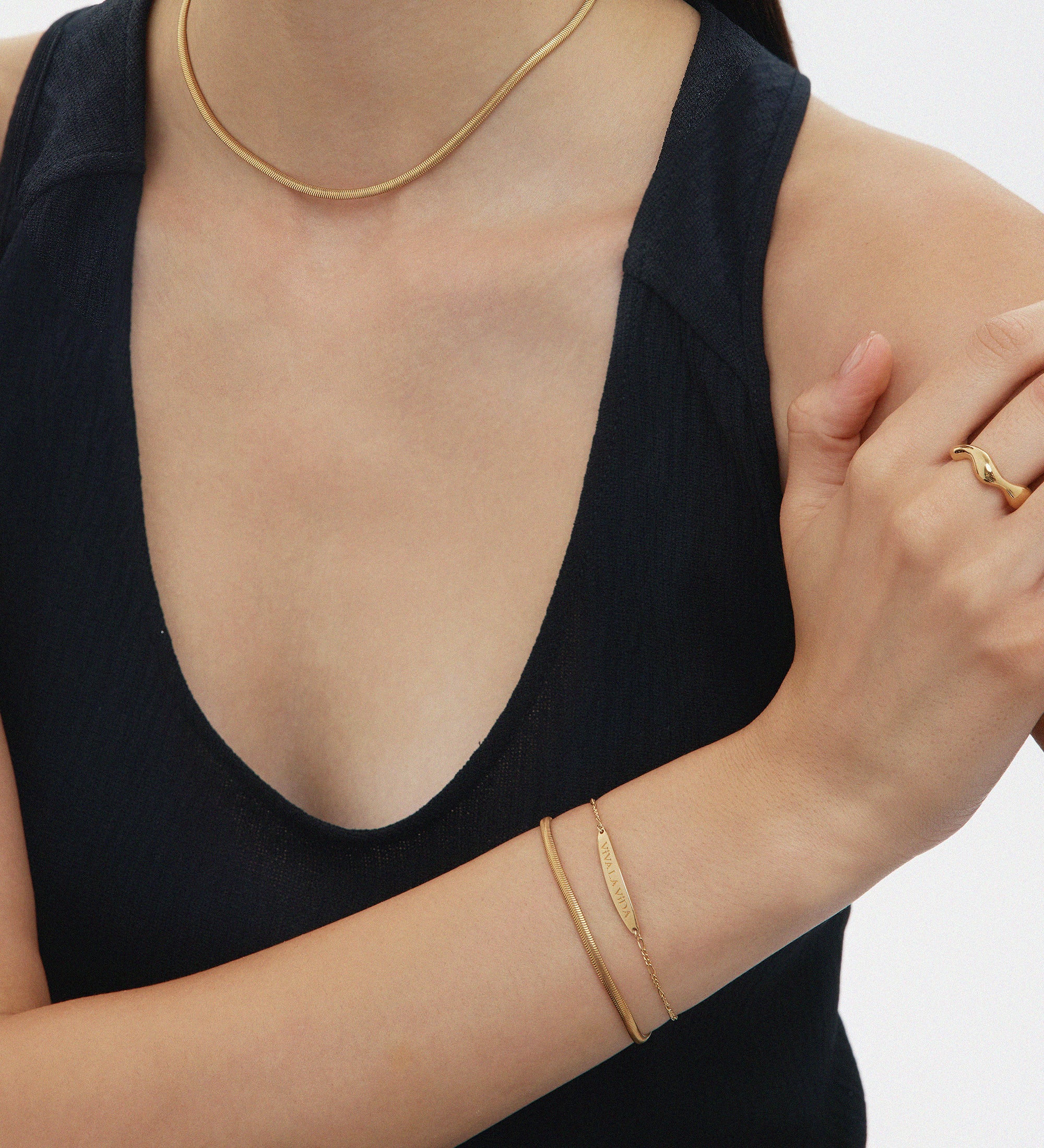 A woman in a sleeveless black top wears a thin gold choker, a simple gold bracelet, a gold ring, and the Estelle Herringbone Chain Necklace in Gold, with her right arm folded across her body.