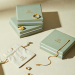 Three sage green VANCII jewelry boxes with gold jewelry—rings, a bracelet, and Judith Hexagon Angular Huggies in Gold—are displayed on a beige surface next to a cream drawstring pouch featuring the VANCII logo.