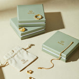 Three sage green VANCII jewelry boxes with gold jewelry—rings, a bracelet, and Judith Hexagon Angular Huggies in Gold—are displayed on a beige surface next to a cream drawstring pouch featuring the VANCII logo.