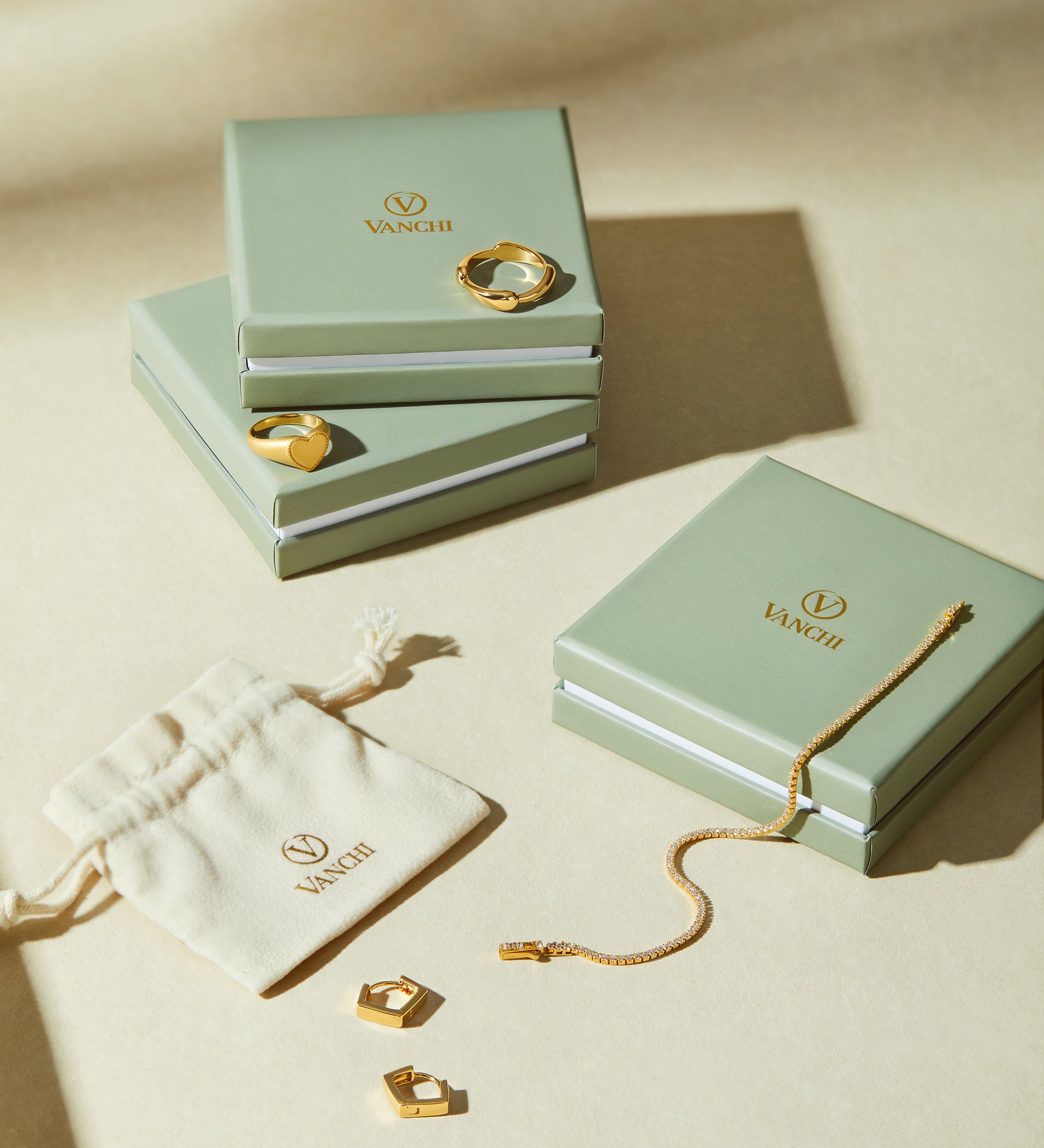 Three sage green VANCII jewelry boxes with gold jewelry—rings, a bracelet, and Judith Hexagon Angular Huggies in Gold—are displayed on a beige surface next to a cream drawstring pouch featuring the VANCII logo.