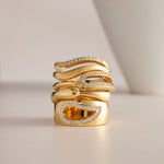 The Colette Link Droop Ring in Gold features a wavy, modern design with gold plating and sparkling diamonds. A rectangular amber gemstone accents the statement ring, all displayed on a neutral background with a white curved object nearby.