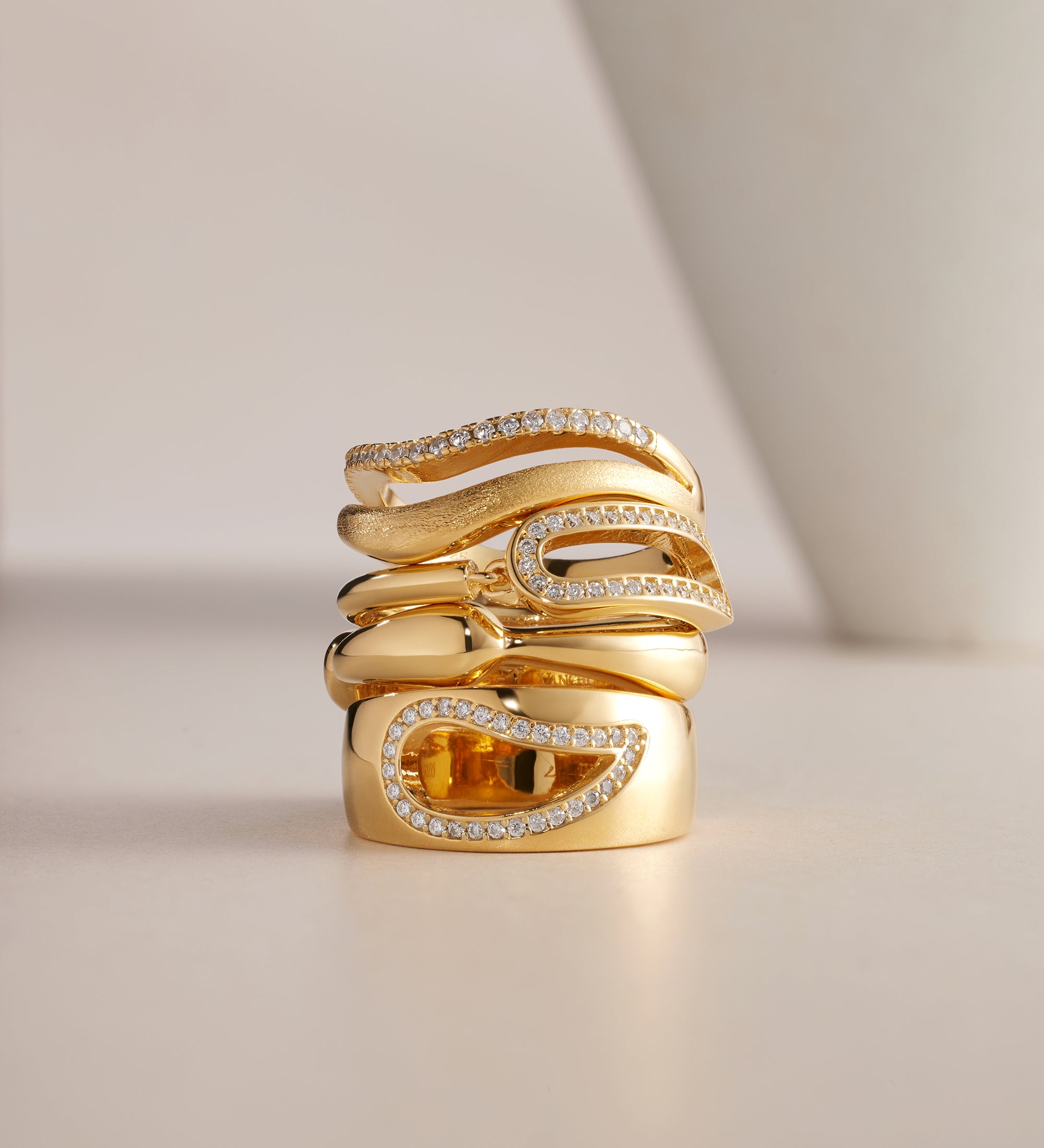 The Colette Link Droop Ring in Gold features a wavy, modern design with gold plating and sparkling diamonds. A rectangular amber gemstone accents the statement ring, all displayed on a neutral background with a white curved object nearby.