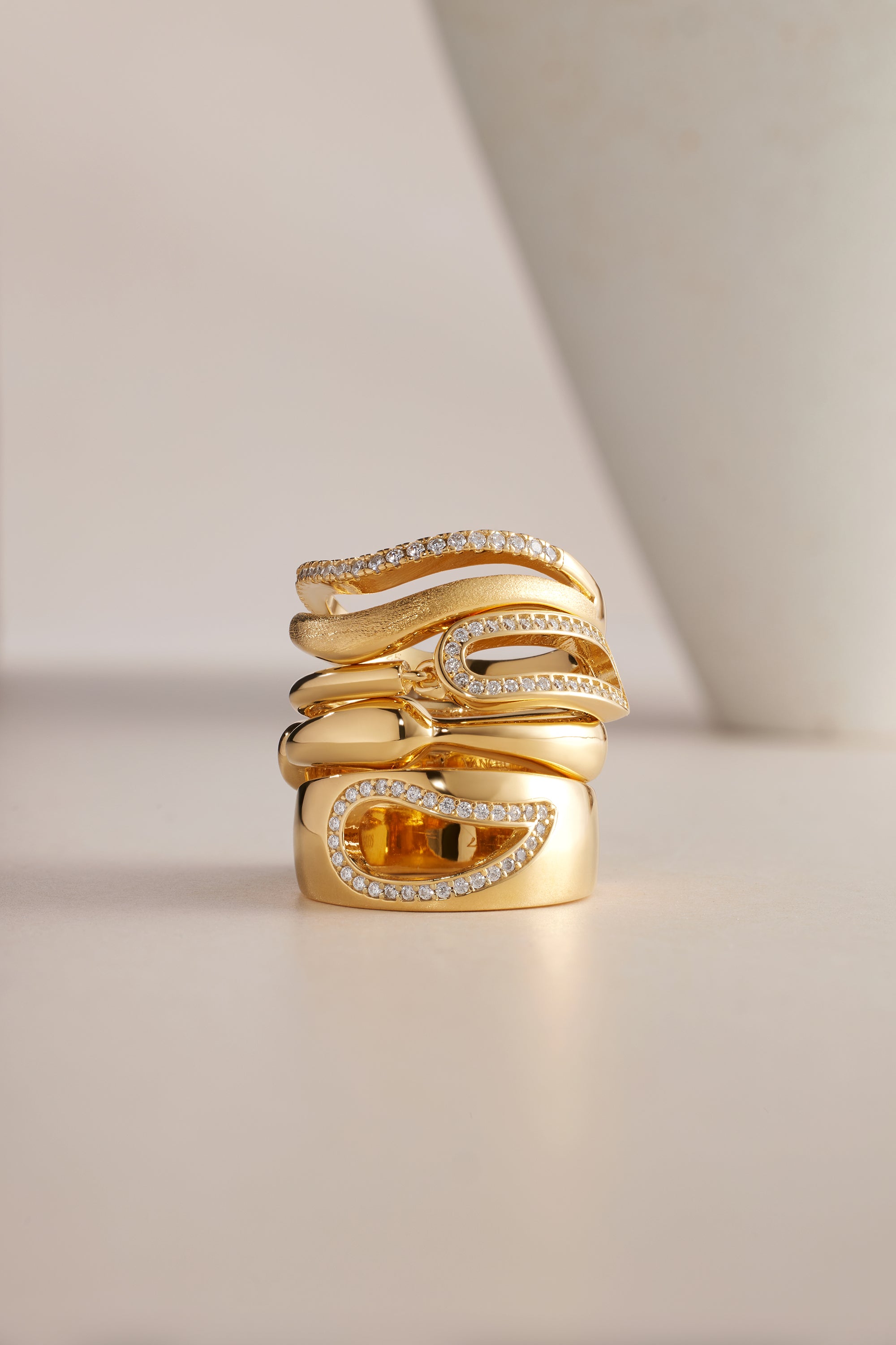 The Colette Link Droop Ring in Gold features a wavy, modern design with gold plating and sparkling diamonds. A rectangular amber gemstone accents the statement ring, all displayed on a neutral background with a white curved object nearby.