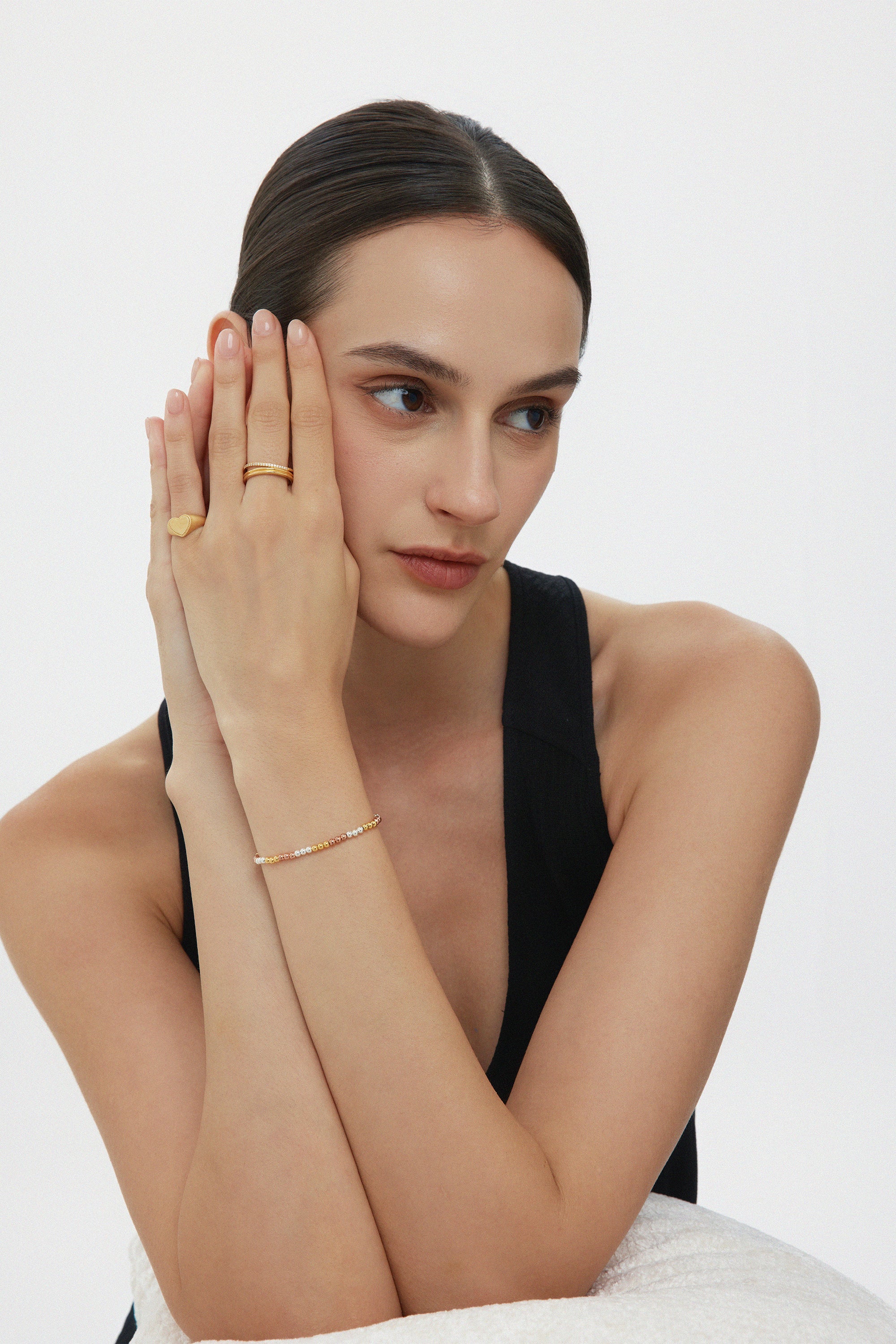 A woman with dark hair in a low bun wears a sleeveless black top and the Noor Pavé Triple Band Ring in Gold, resting her head on her hands and gazing thoughtfully to the side against a plain light background.