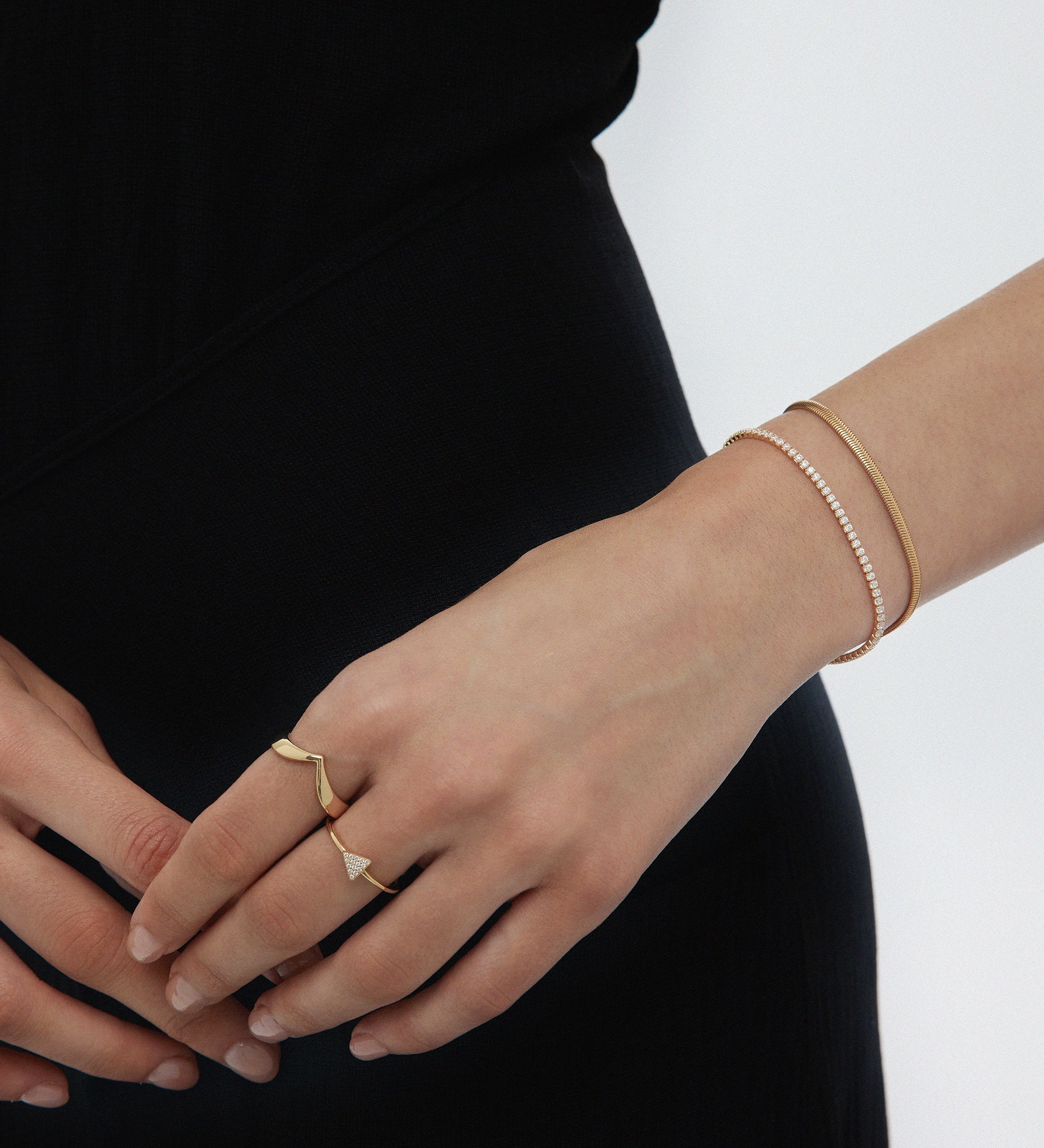 A person in black showcases their left hand with the Veda Plain Chevron Ring in Gold, plus two gold bracelets, against a plain white background.