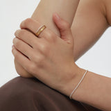 A person with light skin rests their hand on their arm, wearing the Noor Pavé Triple Band Ring in Gold and a delicate diamond bracelet. Theyre dressed in a dark brown garment against a plain, light-colored background.