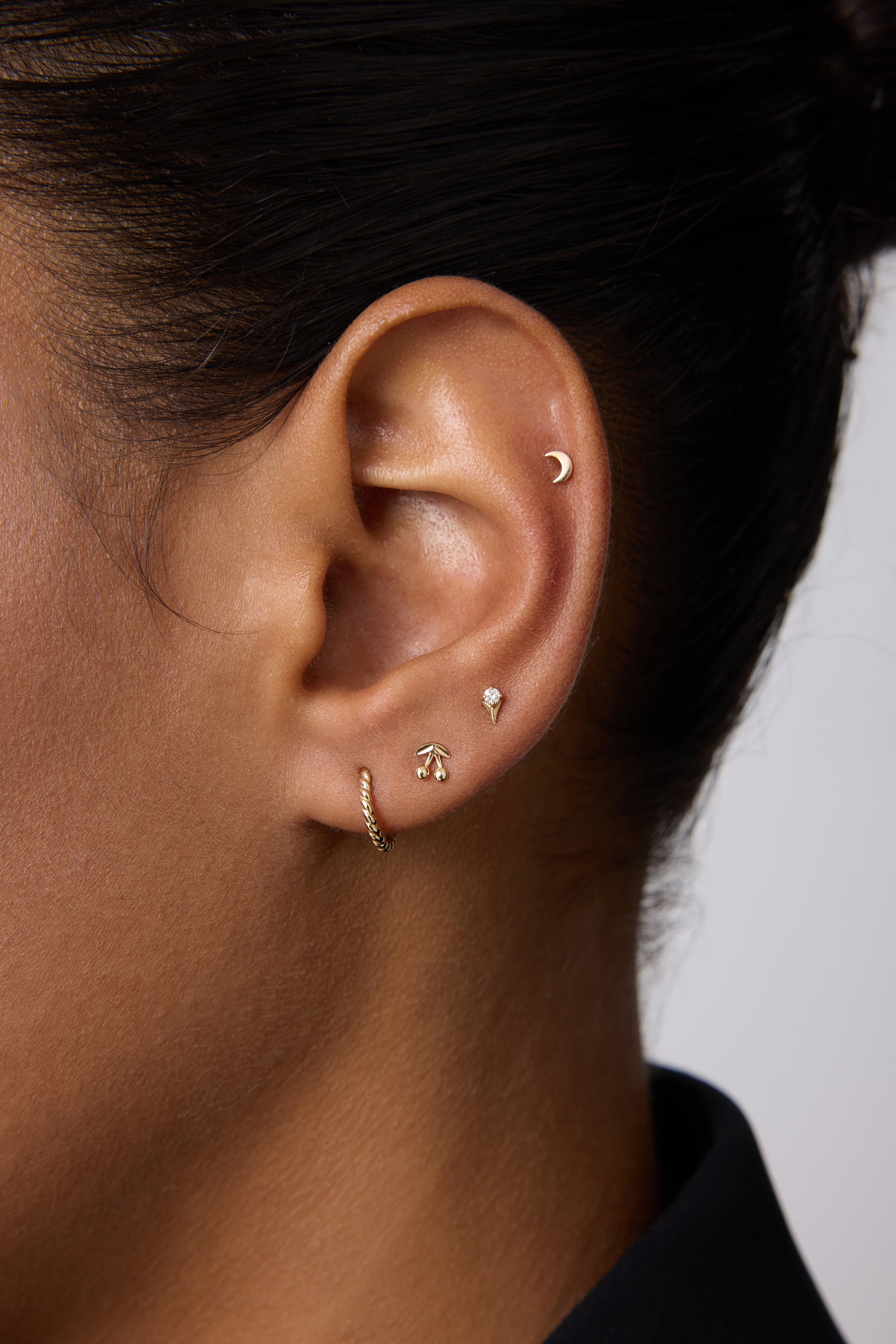 A close-up of an ear with four gold earrings: a crescent moon stud in the upper cartilage, 14K Recycled Solid Yellow Gold Rope Huggie Earrings in the lobe, and two small shaped studs above the huggie.