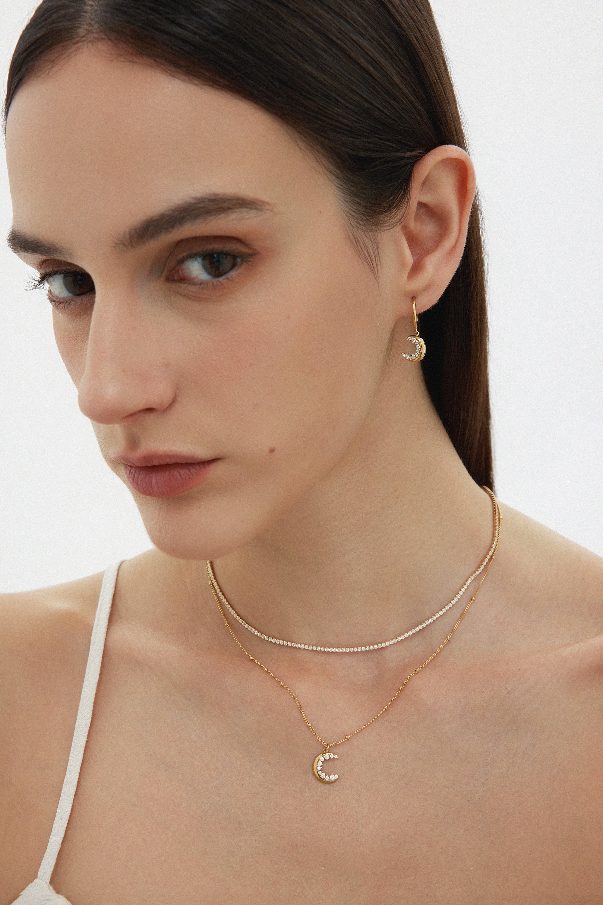 A woman with straight brown hair wears a white top, gold hoop earrings with a crescent moon charm, and a layered silver Alba Tennis Necklace—one strand features a matching crescent moon pendant. She has soft makeup and a neutral expression.