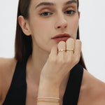 A woman with straight brown hair and natural makeup rests her chin on her hand, showcasing the Veda Plain Chevron Ring in Gold. She wears a sleeveless black top and looks directly at the camera.