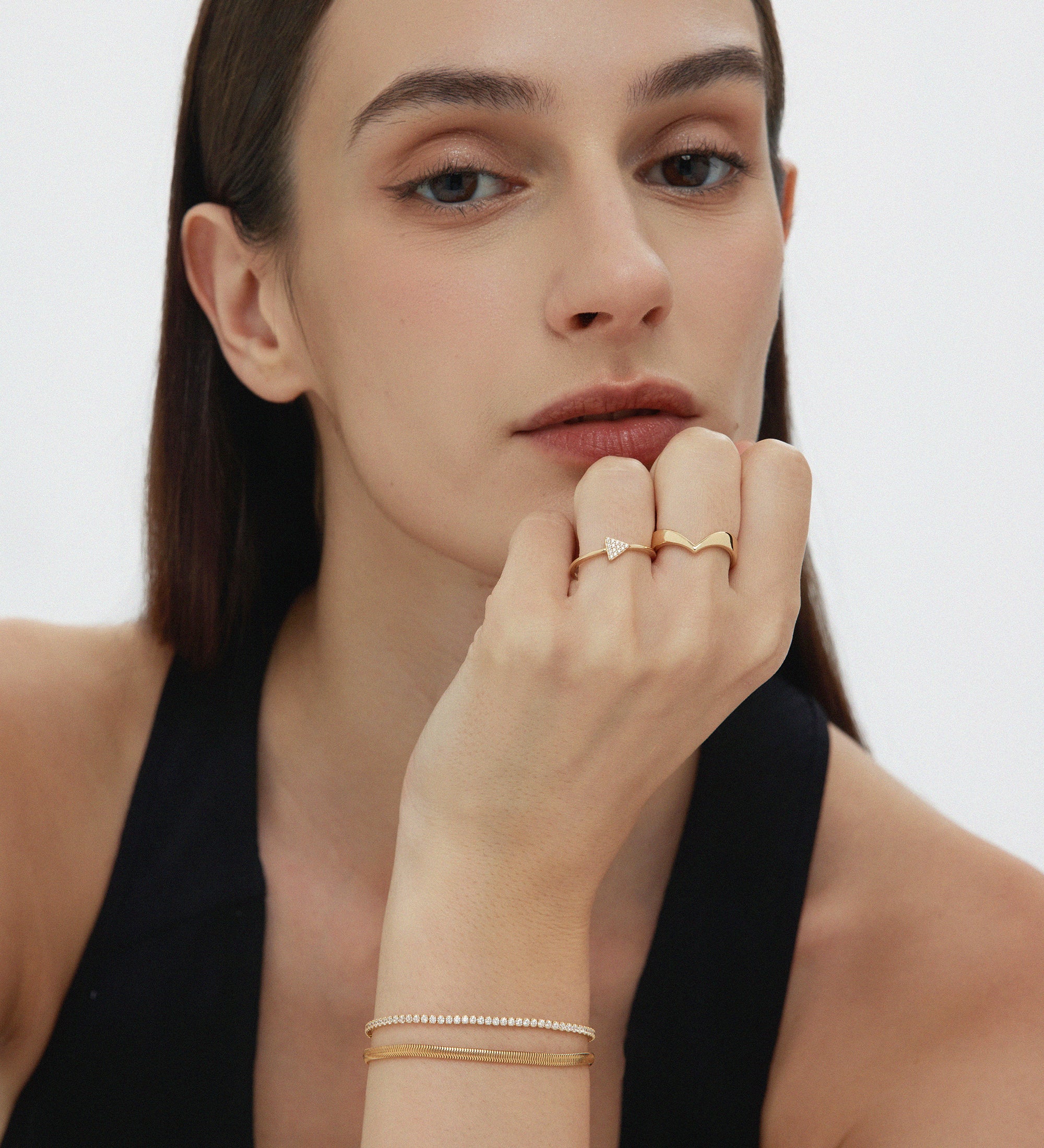 A woman with straight brown hair and natural makeup rests her chin on her hand, showcasing the Veda Plain Chevron Ring in Gold. She wears a sleeveless black top and looks directly at the camera.