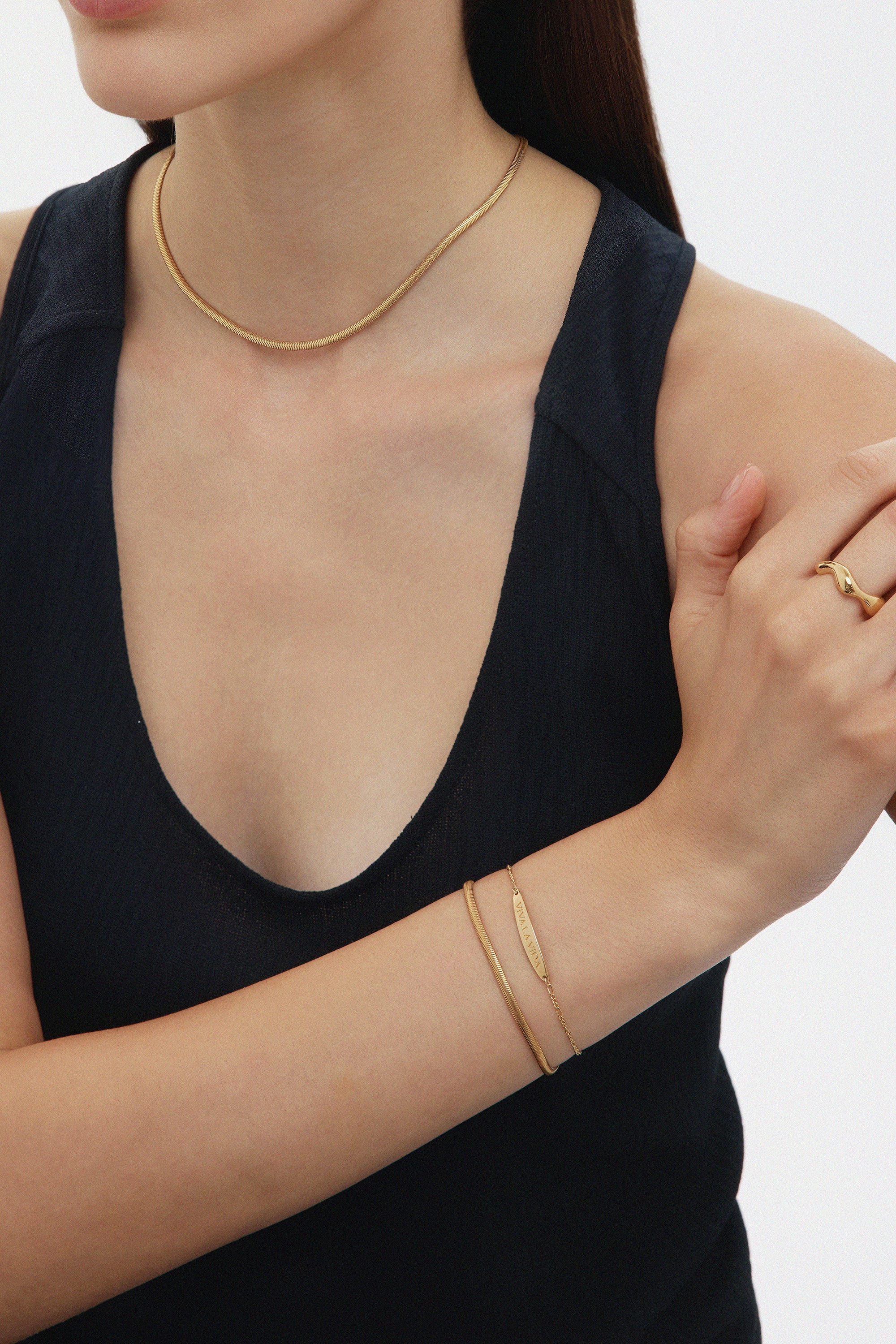 A woman in a sleeveless black top wears a gold figaro chain, a gold ring, and the Viva La Vida Bar Chain Bracelet in Gold. She crosses one arm over her chest against a plain white background, with her face partially out of frame.