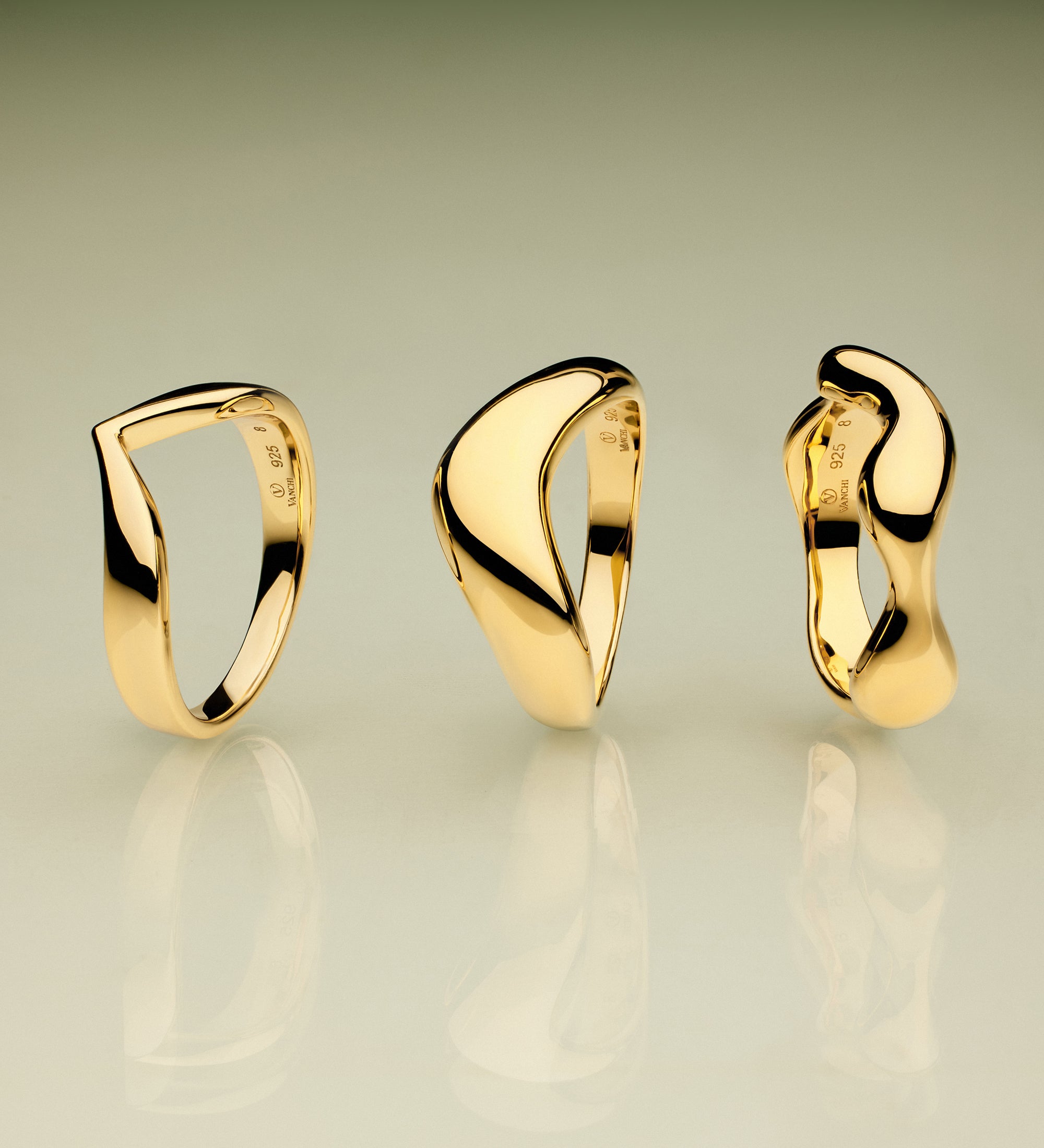 The Vava Plain Chunky Curved Wave Ring in Gold, with its modern sculptural form, stands upright on a reflective surface against a green-to-beige gradient background—a striking showcase of minimalist jewelry.