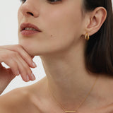 A woman with straight dark hair wears minimalist jewellery, featuring the Regina Horizontal Bar Pendant Necklace in Gold and small drop earrings, as she poses with her chin on her hand against a plain light background.