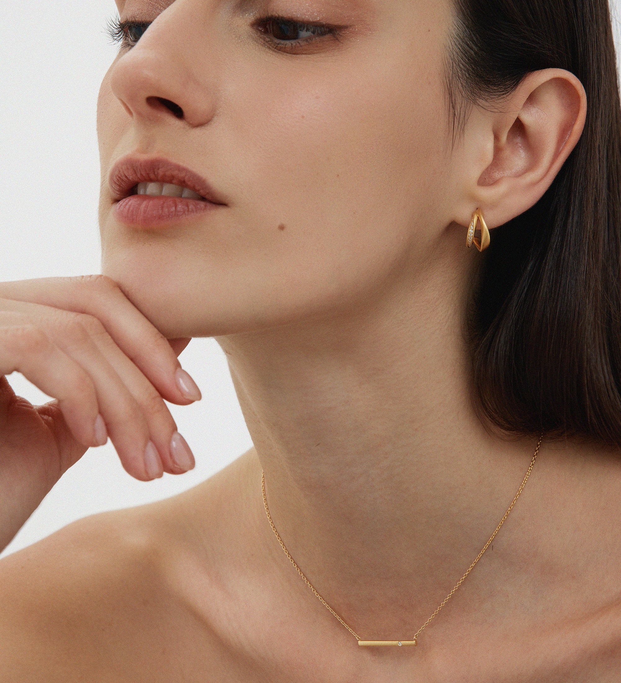 A woman with straight dark hair wears minimalist jewellery, featuring the Regina Horizontal Bar Pendant Necklace in Gold and small drop earrings, as she poses with her chin on her hand against a plain light background.