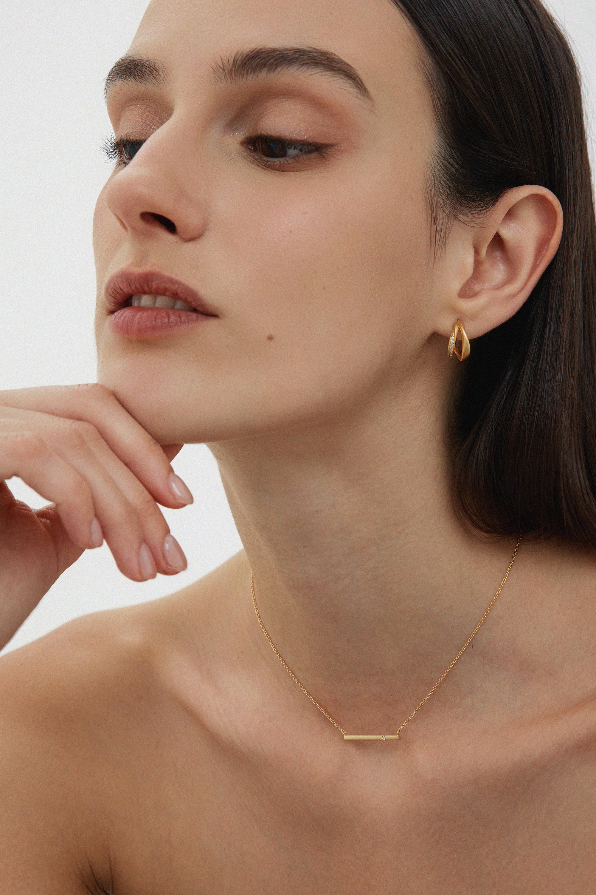 A woman with straight dark hair wears minimalist jewellery, featuring the Regina Horizontal Bar Pendant Necklace in Gold and small drop earrings, as she poses with her chin on her hand against a plain light background.