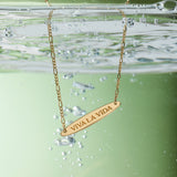 The Viva La Vida Bar Chain Necklace in Gold features VIVA LA VIDA engraved on a partially submerged pendant with bubbles and an adjustable figaro chain, set against a green background.