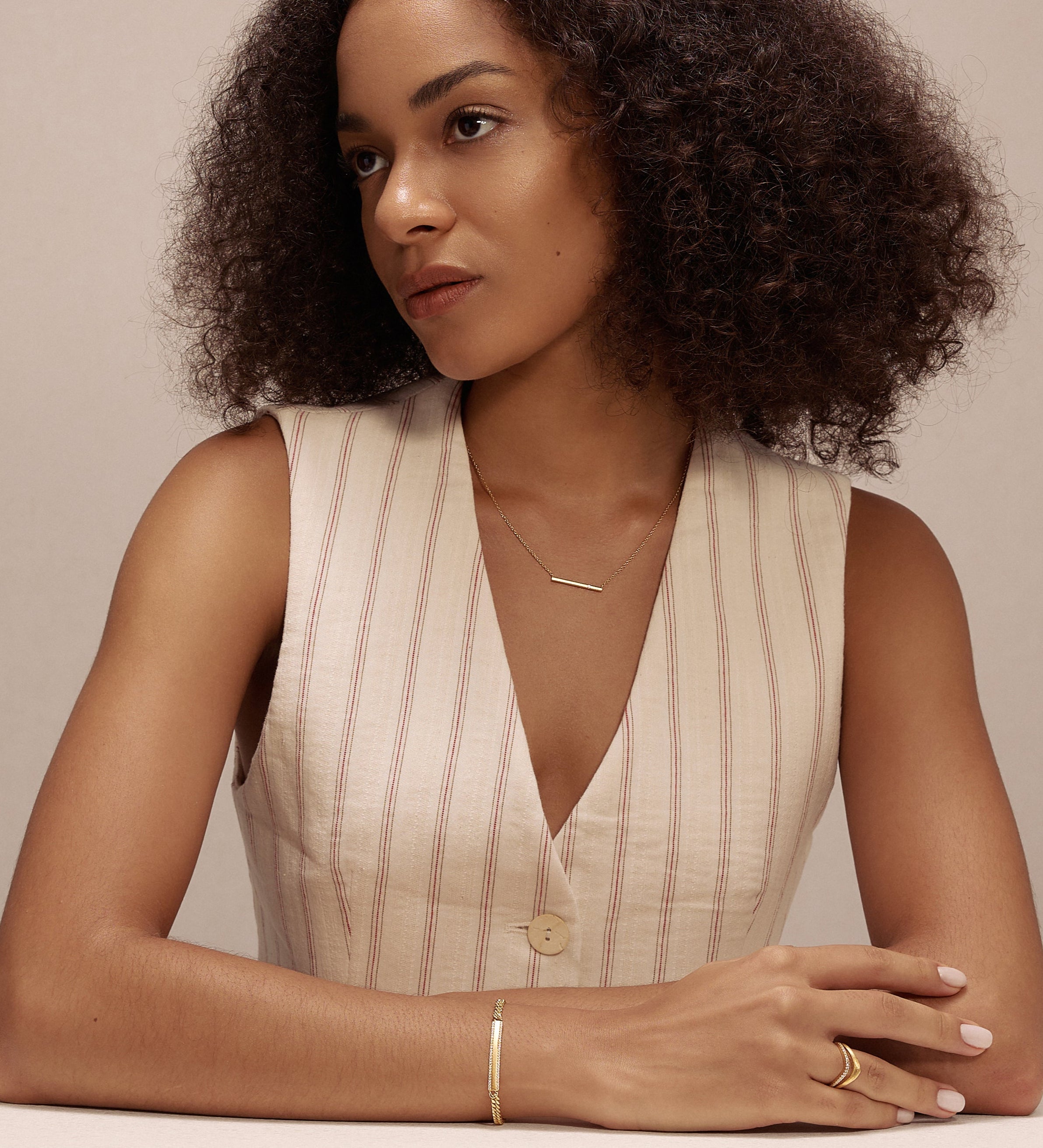 A woman with curly hair, wearing a sleeveless beige vest and the Regina Horizontal Bar Pendant Necklace in Gold, sits at a white table, arms resting on it, gazing thoughtfully to the side against a plain light background.