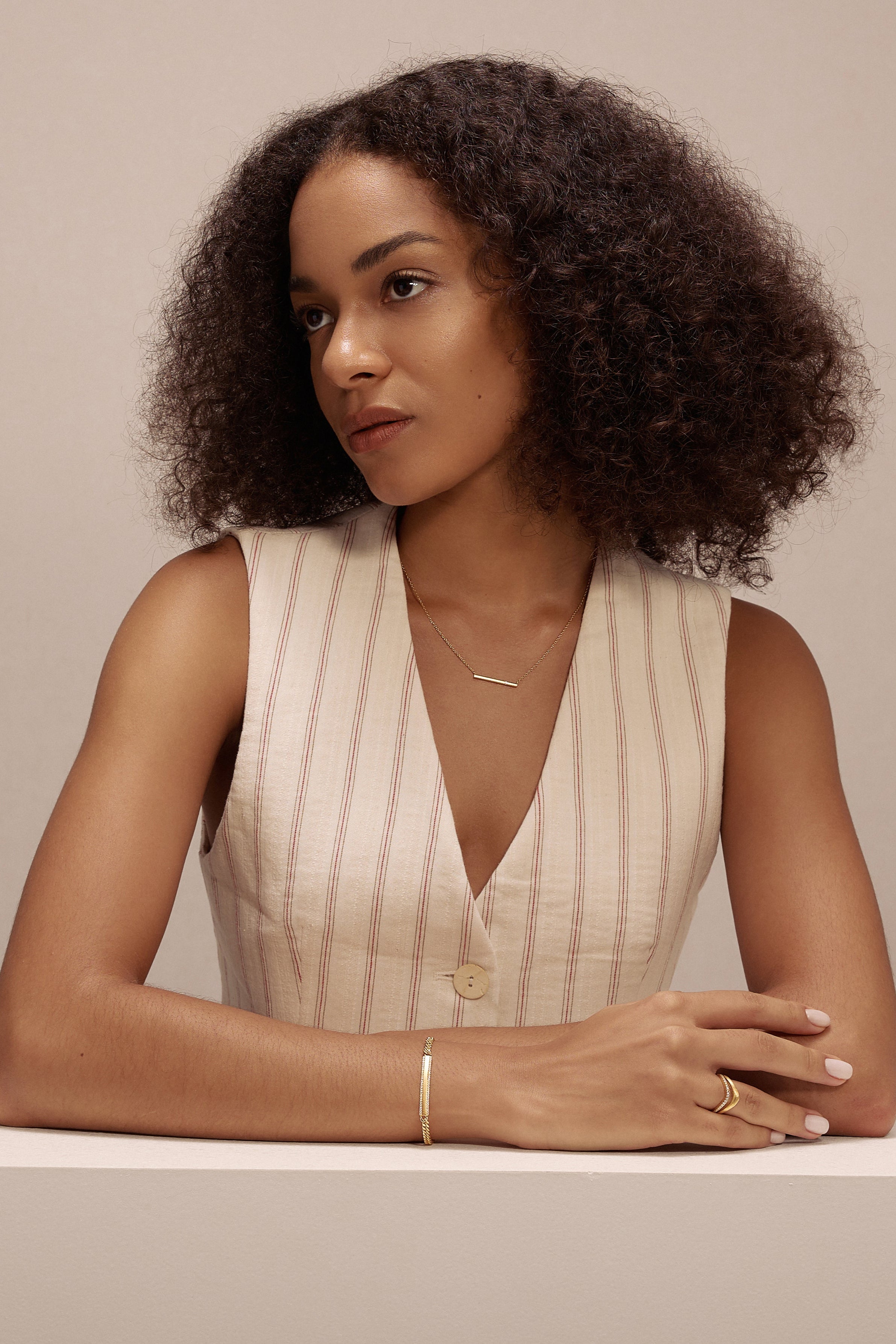 A woman with curly hair, wearing a sleeveless beige vest and the Regina Horizontal Bar Pendant Necklace in Gold, sits at a white table, arms resting on it, gazing thoughtfully to the side against a plain light background.